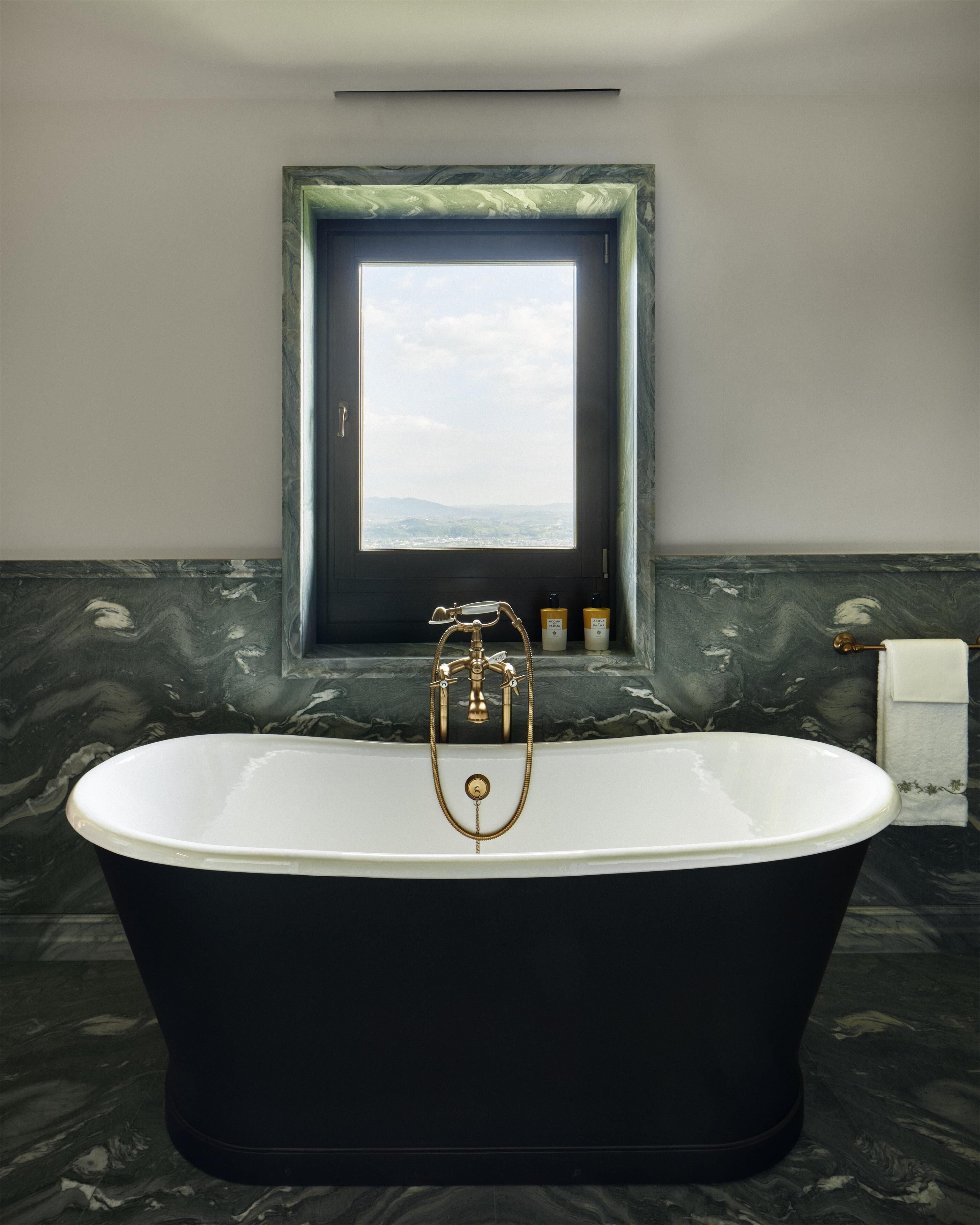 A freestanding bathtub with a gold faucet, a window above it, and a marble wall.