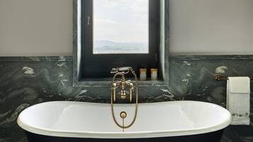 A freestanding bathtub with a gold faucet, a window above it, and a marble wall.