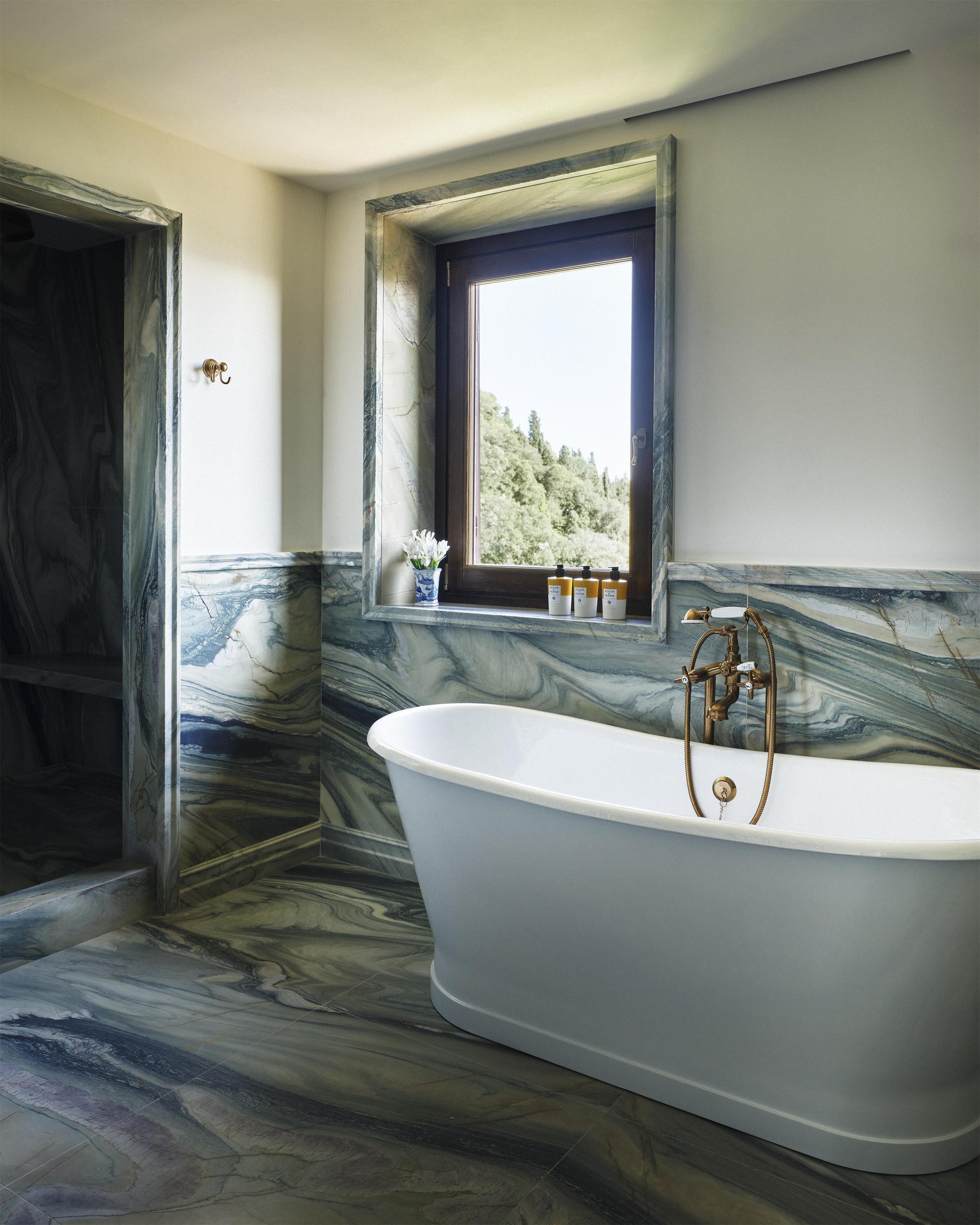 Ein Badezimmer mit einer freistehenden Badewanne, einem Fenster mit Blick auf Bäume und Marmorwänden.
