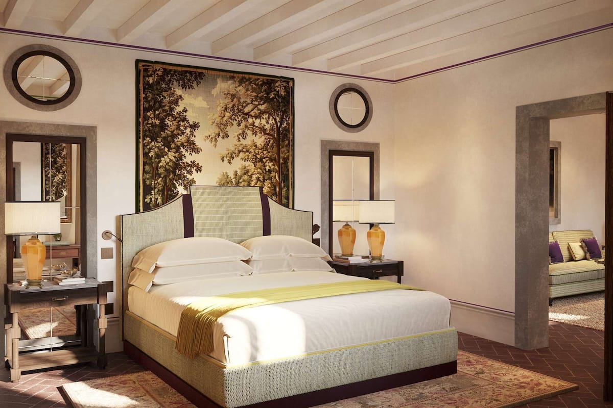 A spacious bedroom with a large bed, two bedside tables with lamps, a large wall tapestry, and a view of a living area.