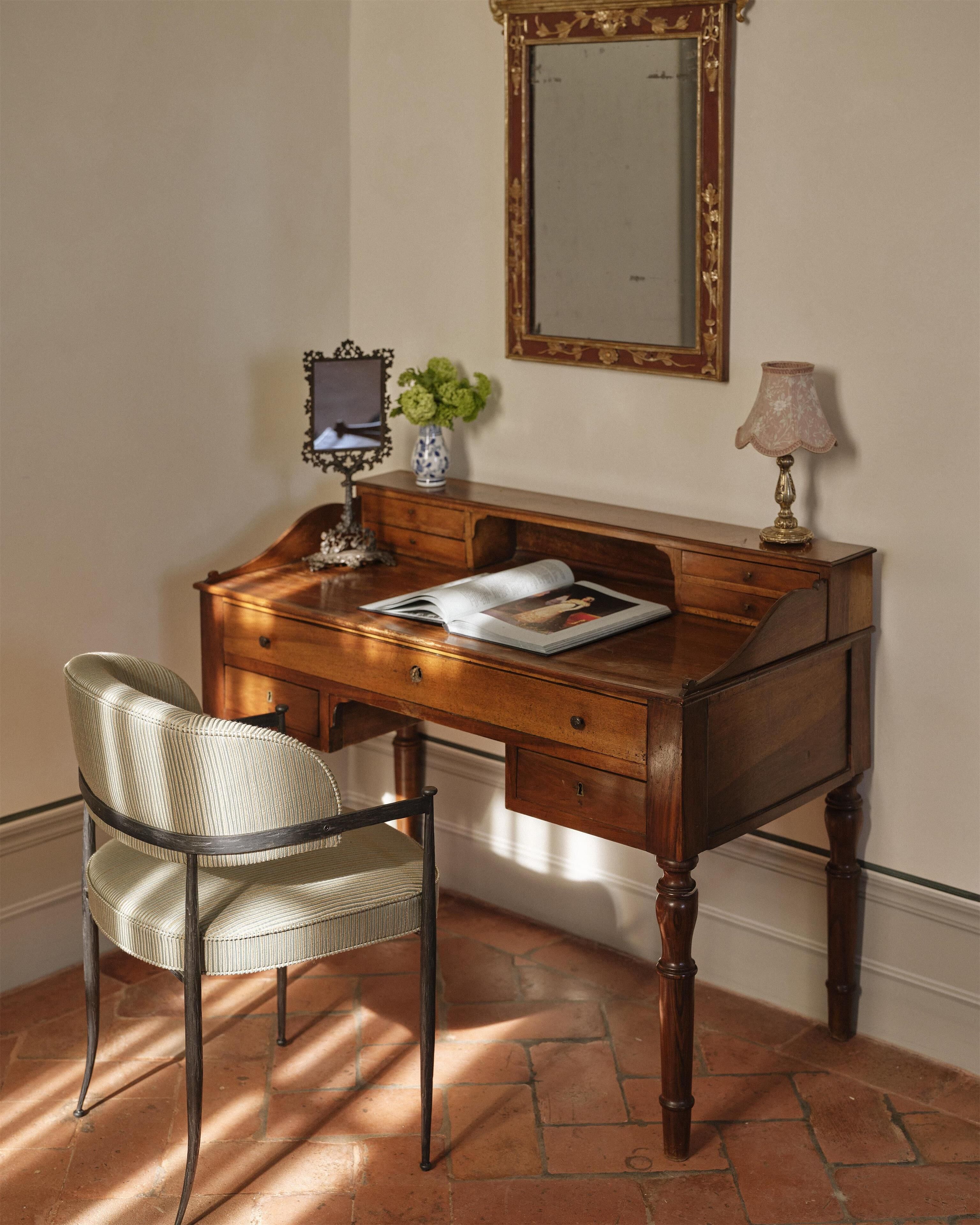 Un escritorio de madera con cajones, una silla, una lámpara, un jarrón con flores y un espejo en la pared.