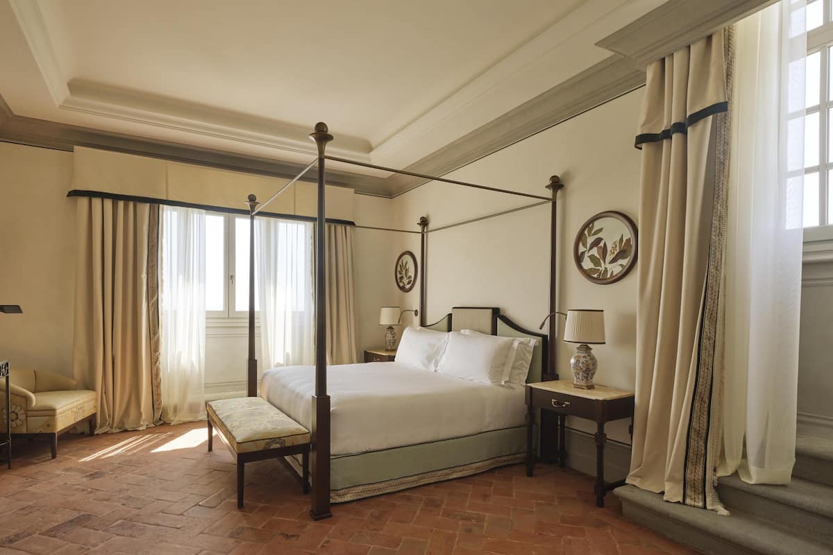 A four-poster bed with a nightstand, a chair, and a window with curtains.