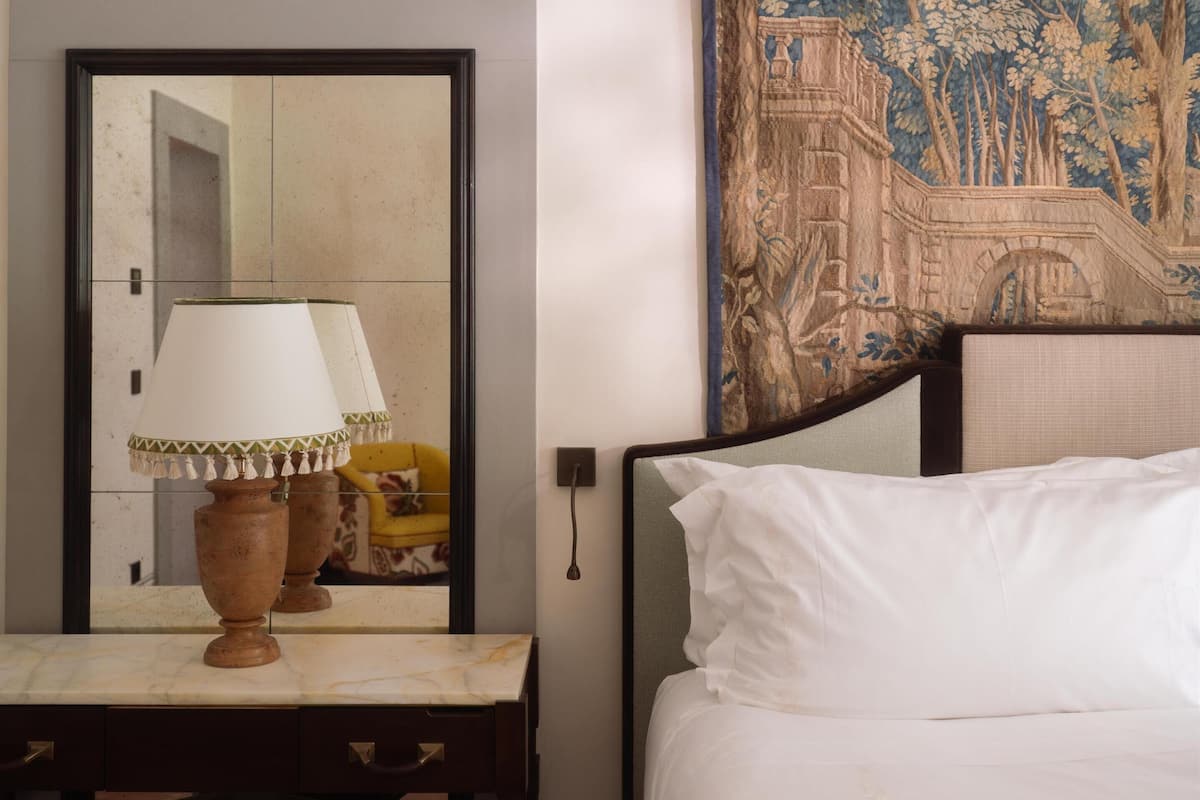 A hotel room with a bed, a round mirror, a table with a lamp, and a tapestry.