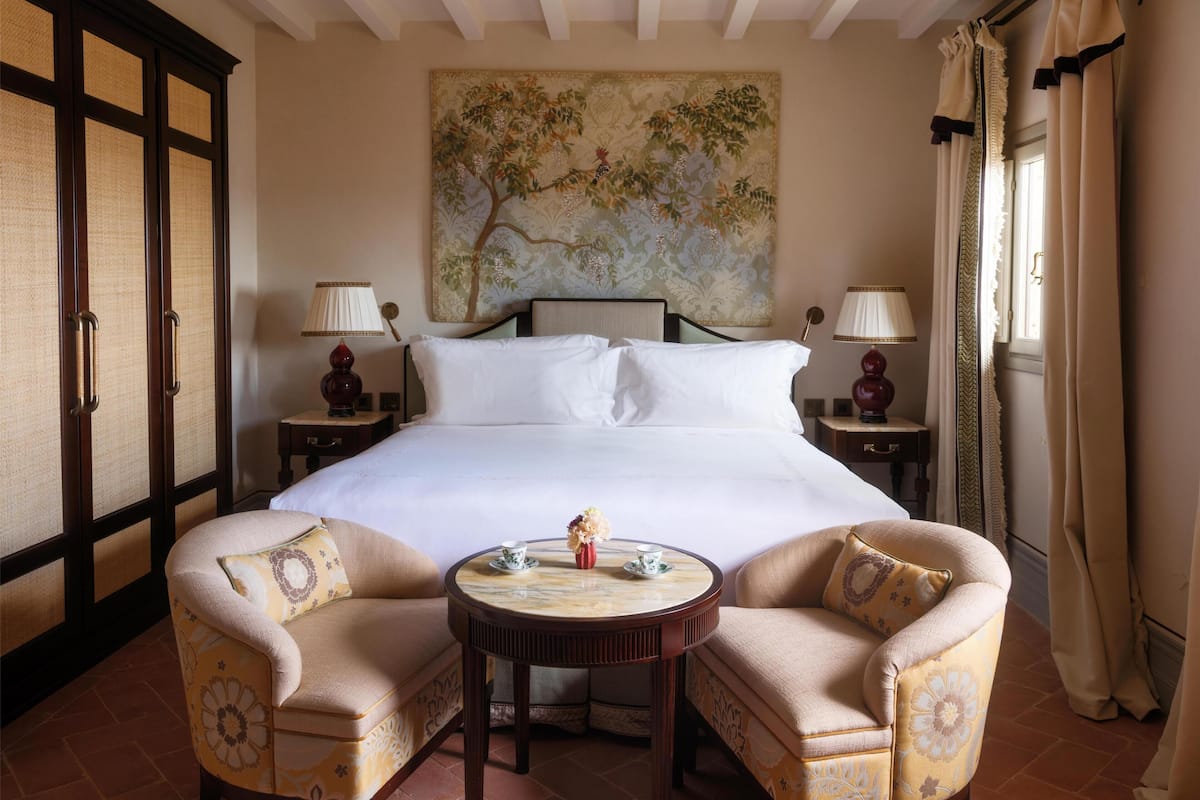 A hotel room with a large bed, two bedside tables, a round table with a vase, two armchairs, and a decorative wall painting.