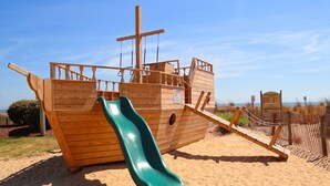 A wooden play structure with a slide and climbing features, set on a sandy beach with a clear sky. - Carousel Resort Hotel & Condominiums (Ocean City)