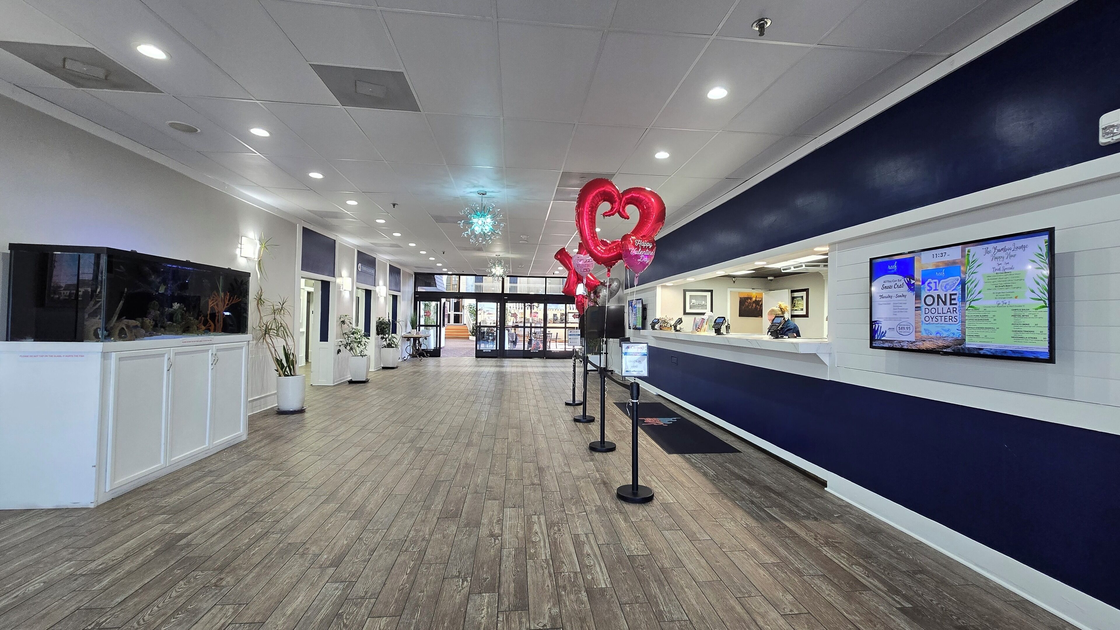 A spacious hallway with wooden flooring, a reception desk, and a large digital screen displaying information.