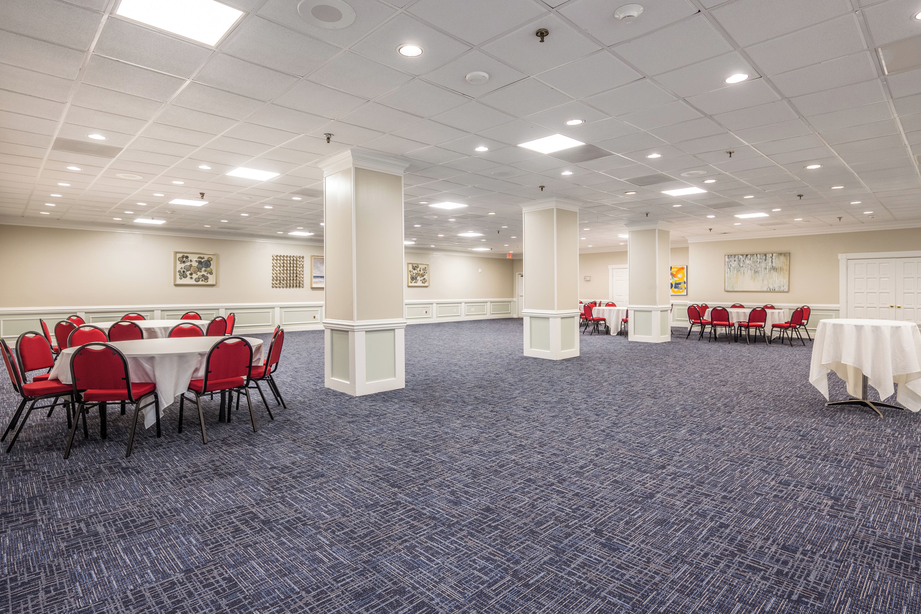 Een ruime congreszaal met meerdere tafels en stoelen, witte zuilen en een kamerbreed tapijt met een patroon.