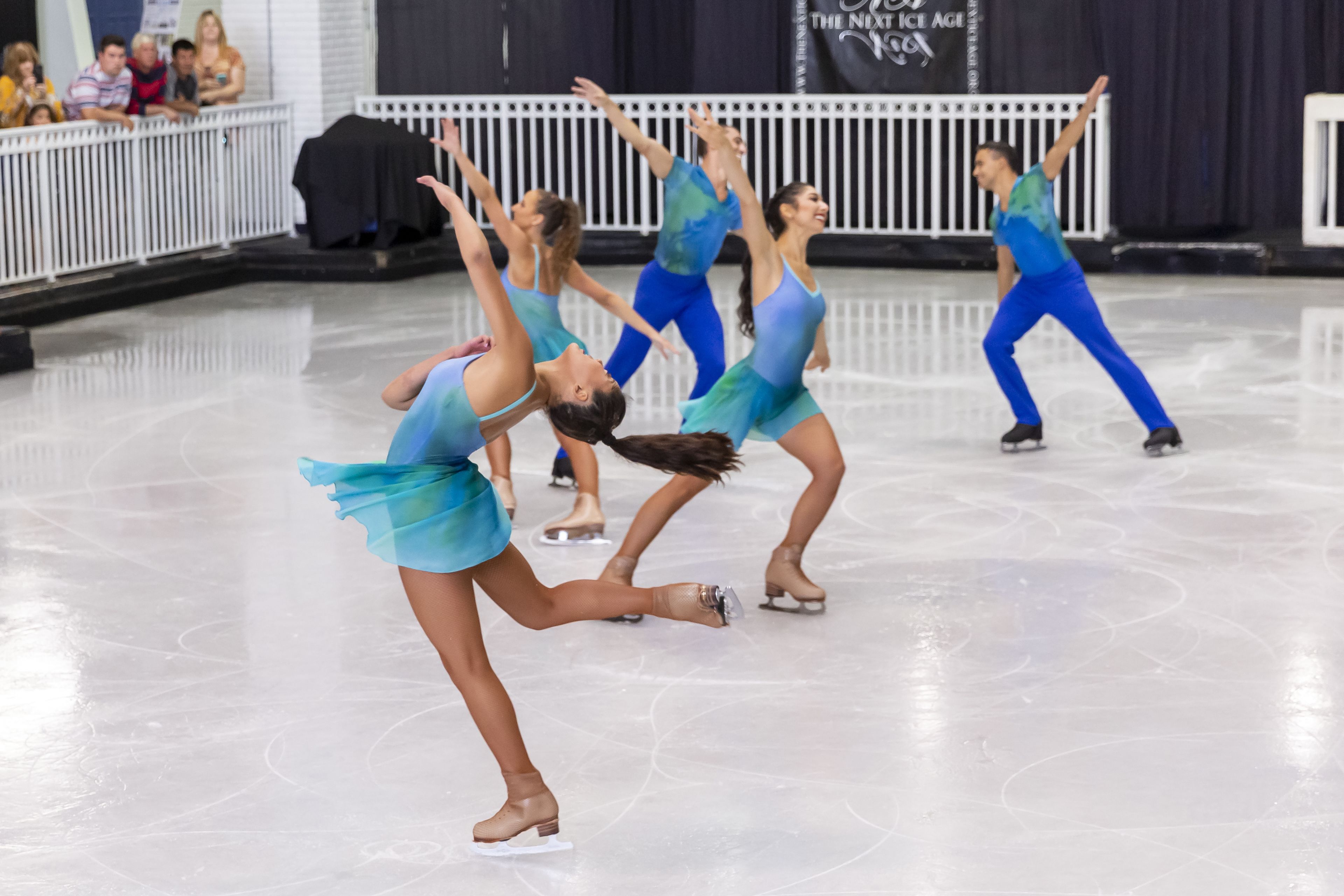 Une séance d’entraînement de patinage artistique, où des patineurs vêtus de costumes bleus et turquoise exécutent des figures sur une patinoire.