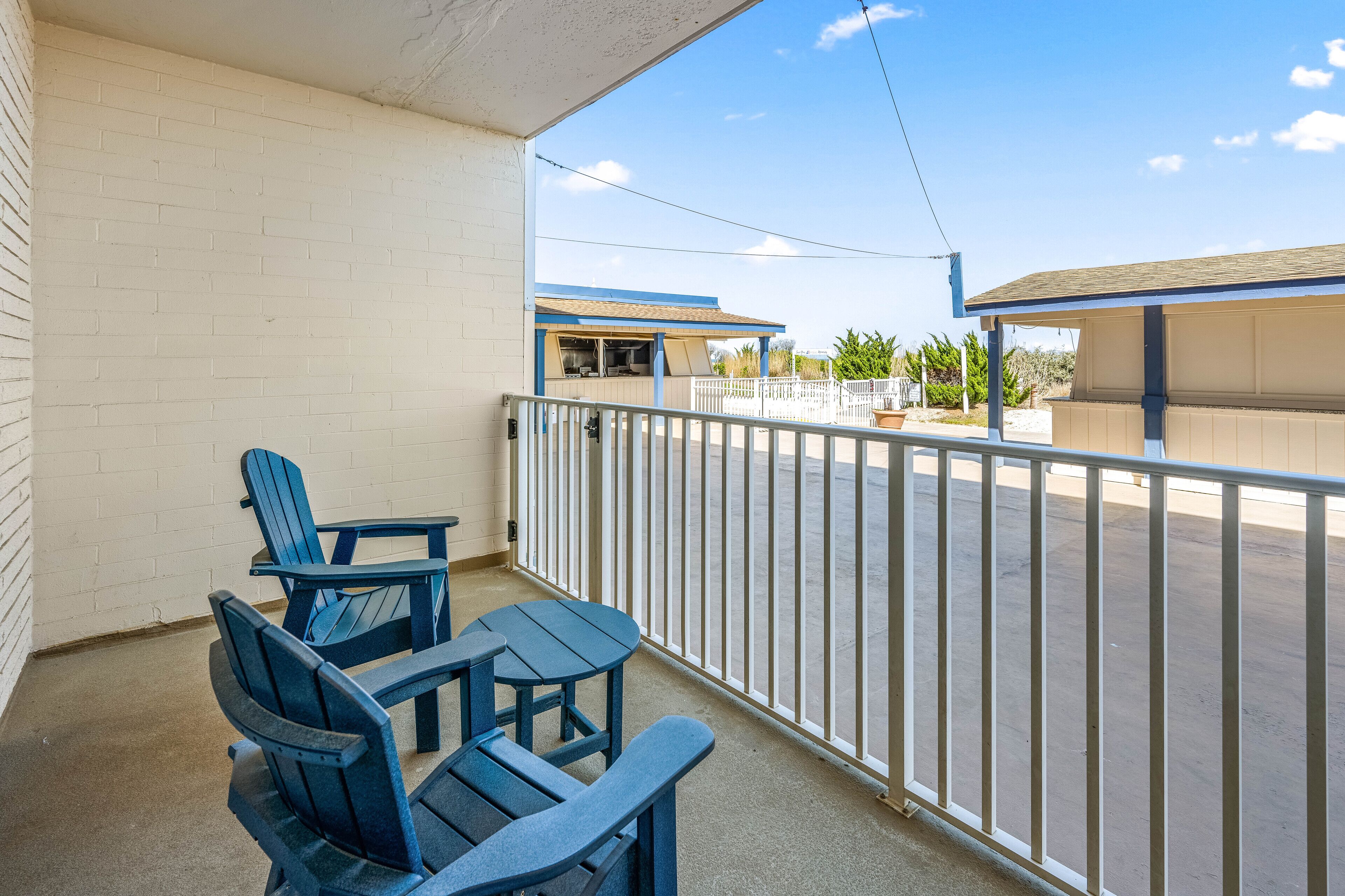 A balcony with blue Adirondack chairs and a table, overlooking a parking lot and buildings.