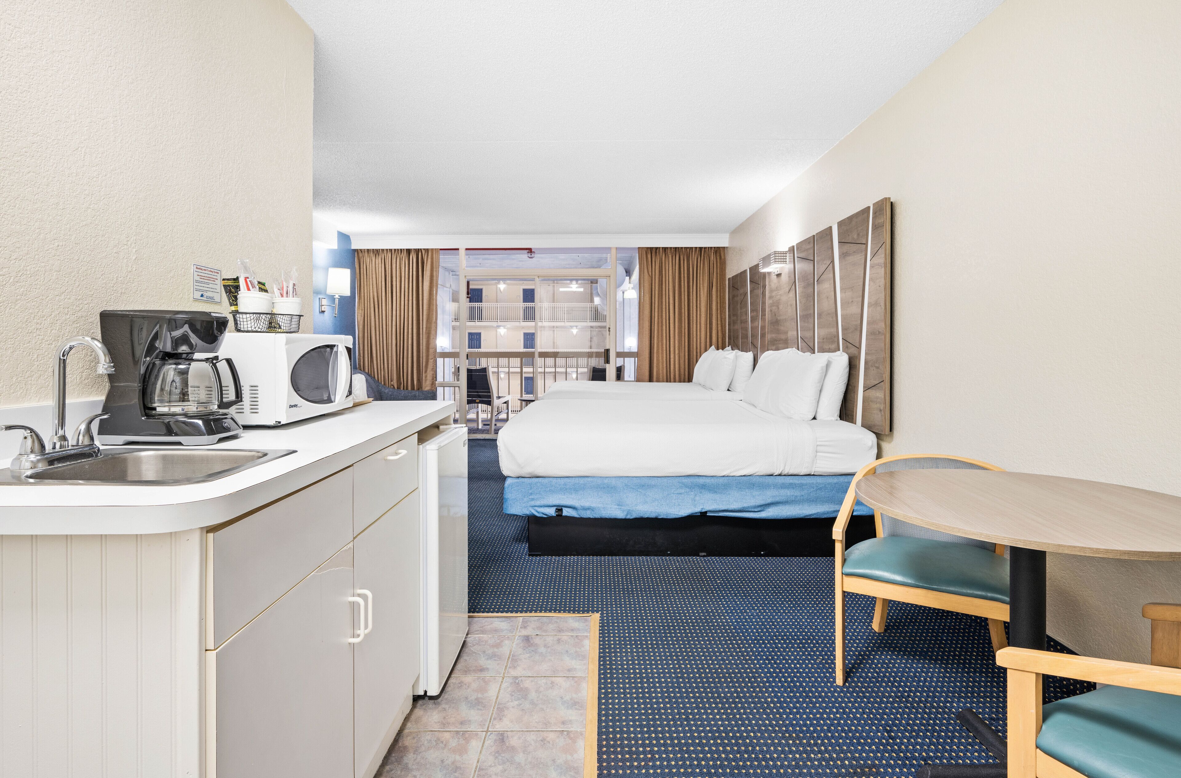 A hotel room with a bed, a small table, a kitchenette, and a view of the building's interior.