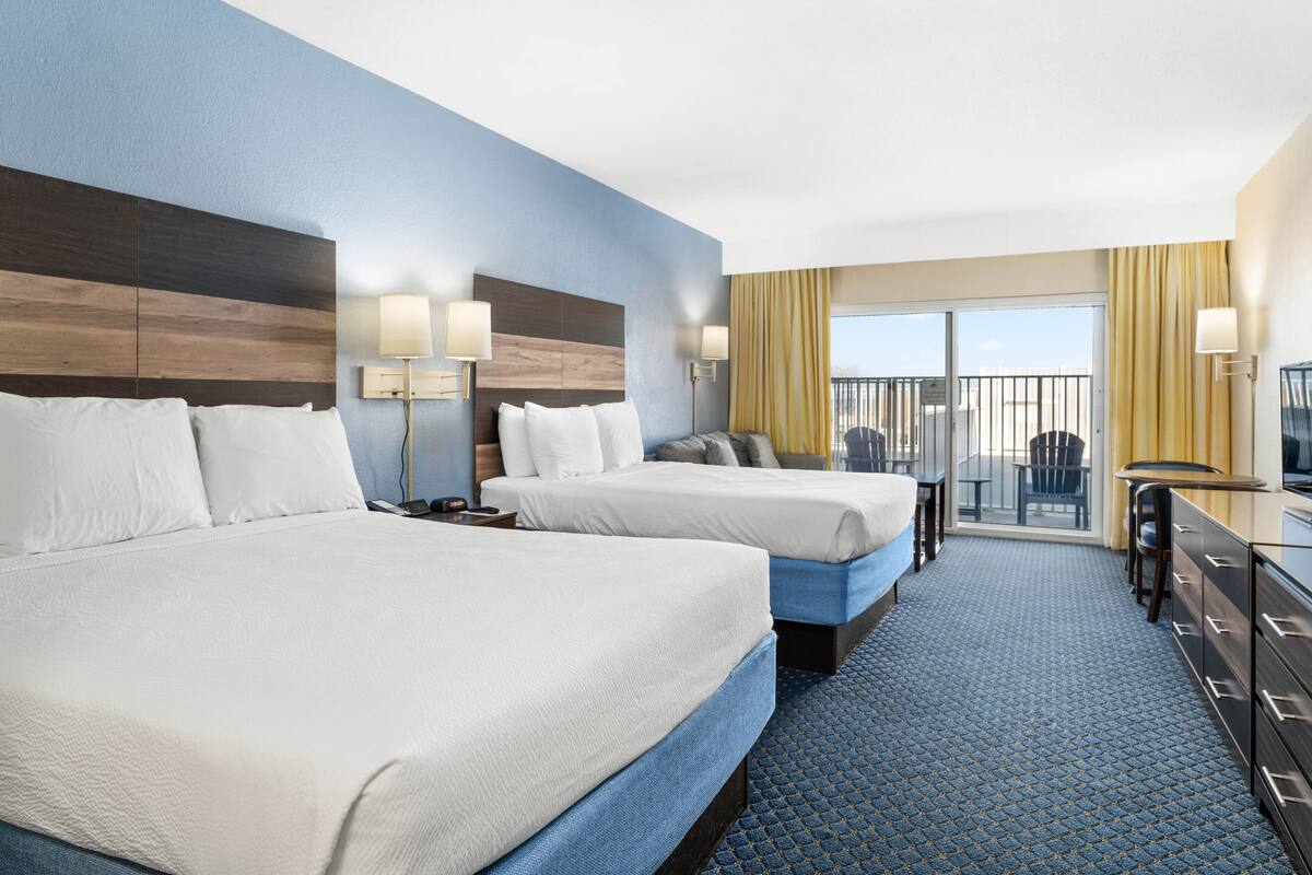 A hotel room with two beds, a desk, and a balcony with yellow curtains.
