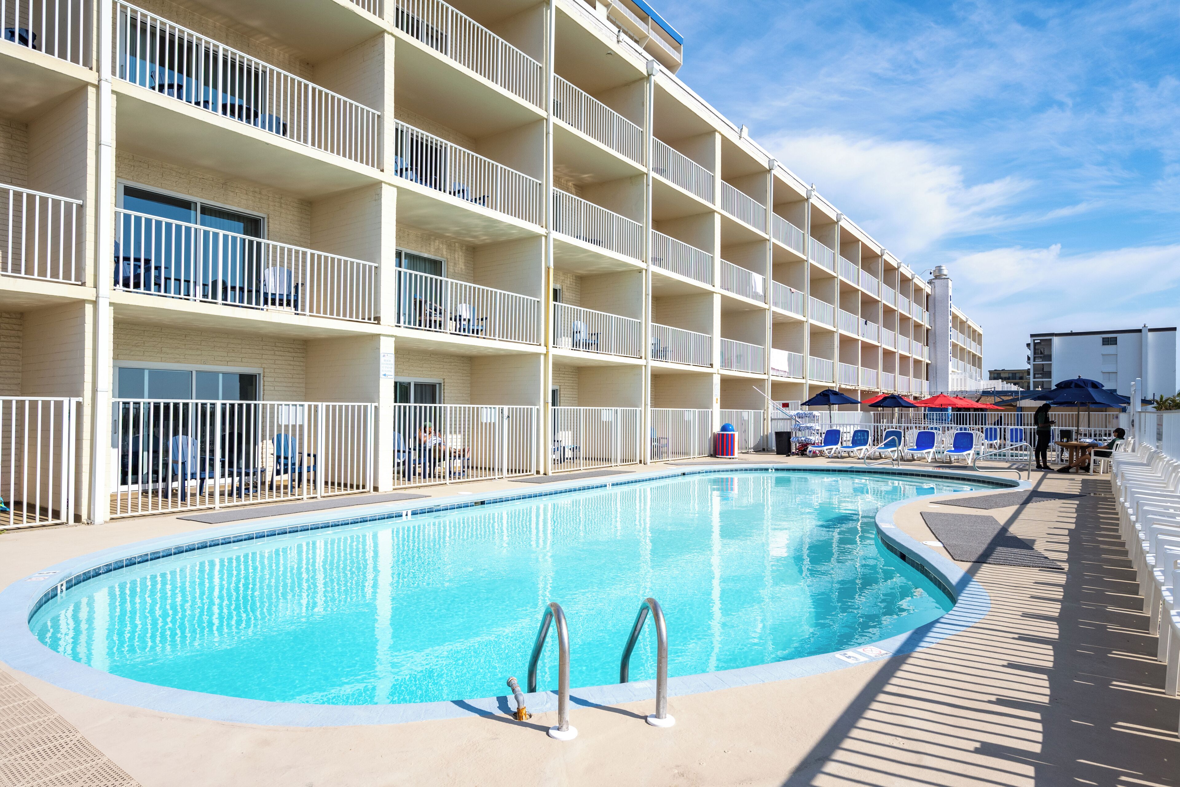 A swimming pool with a deck, surrounded by a multi-story building with balconies and a covered seating area.
