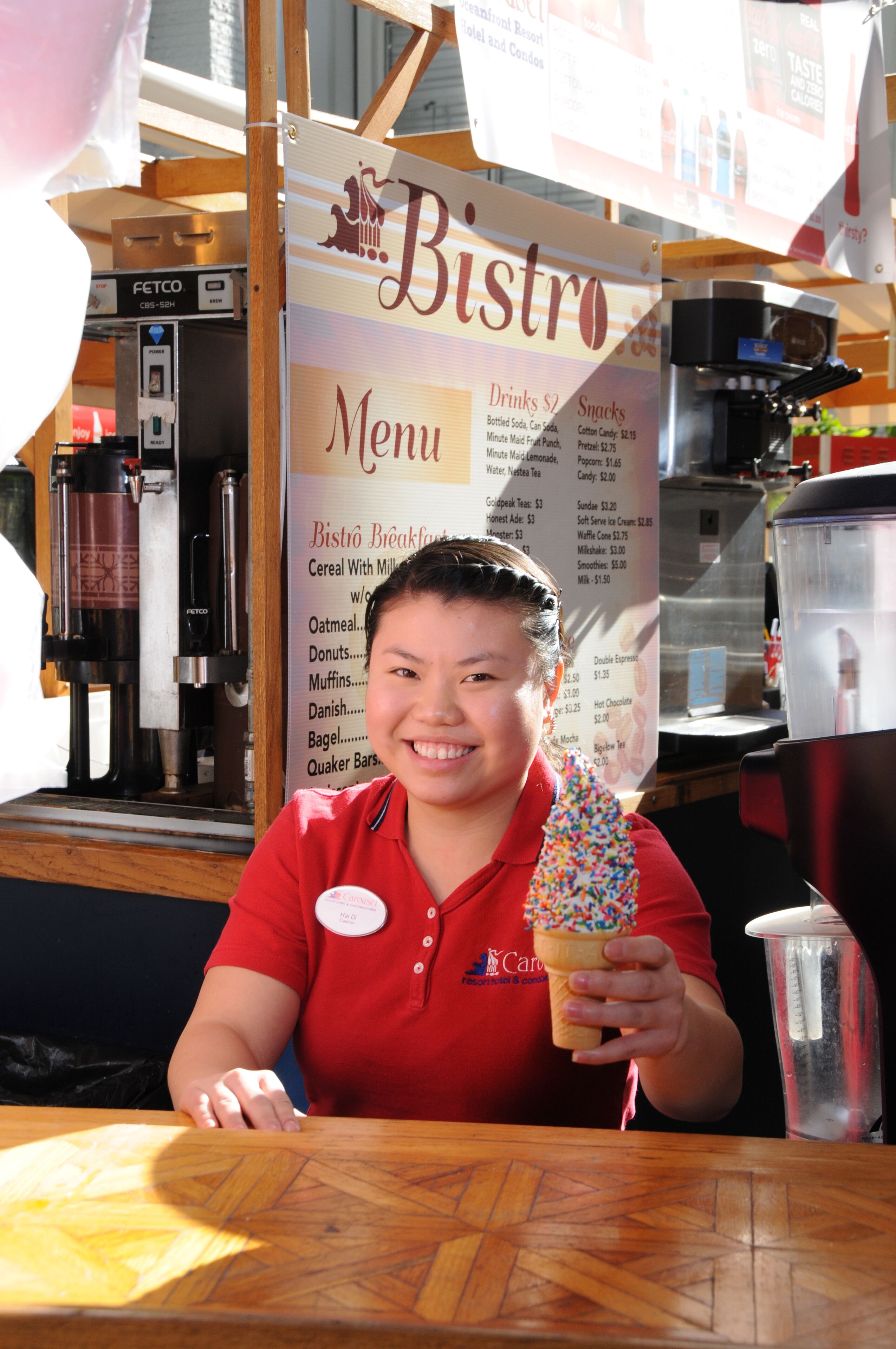 A person in a red shirt holding a cone with sprinkles, sitting at a wooden table with a menu board in the background.