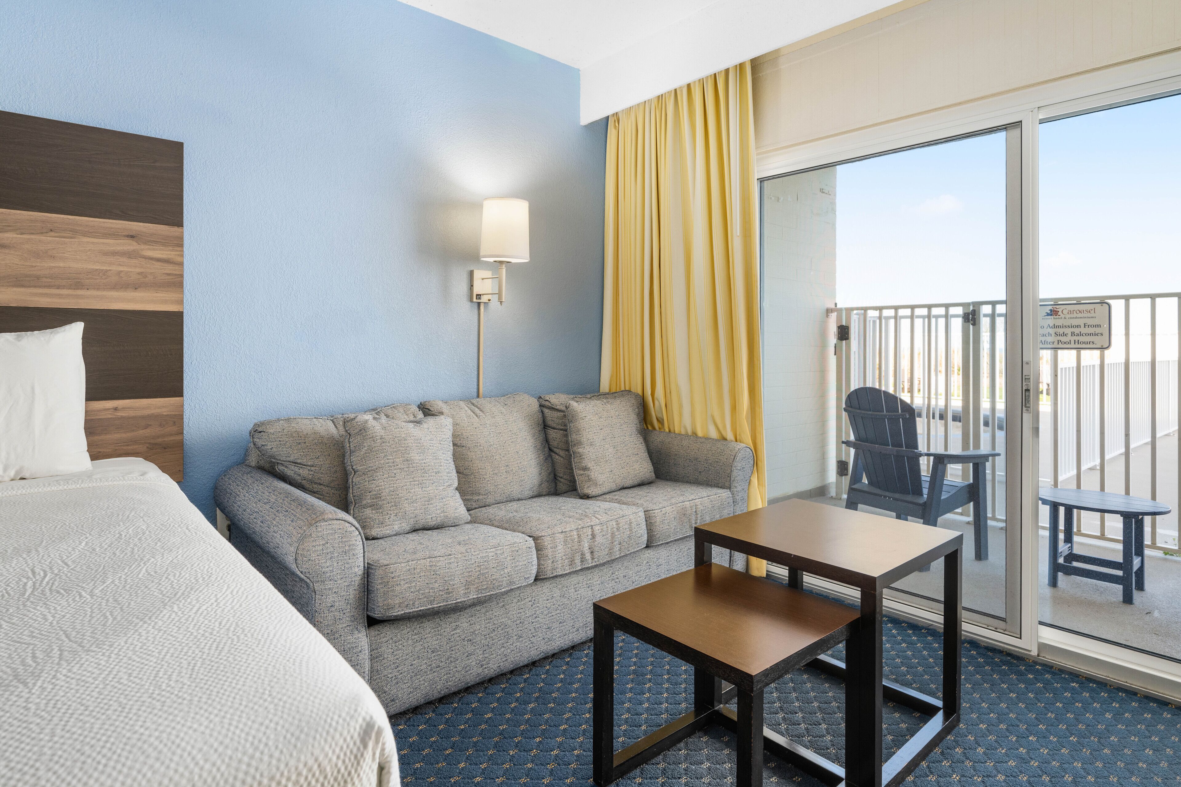 A hotel room with a bed, a grey sofa, a small table, and a balcony with a chair.