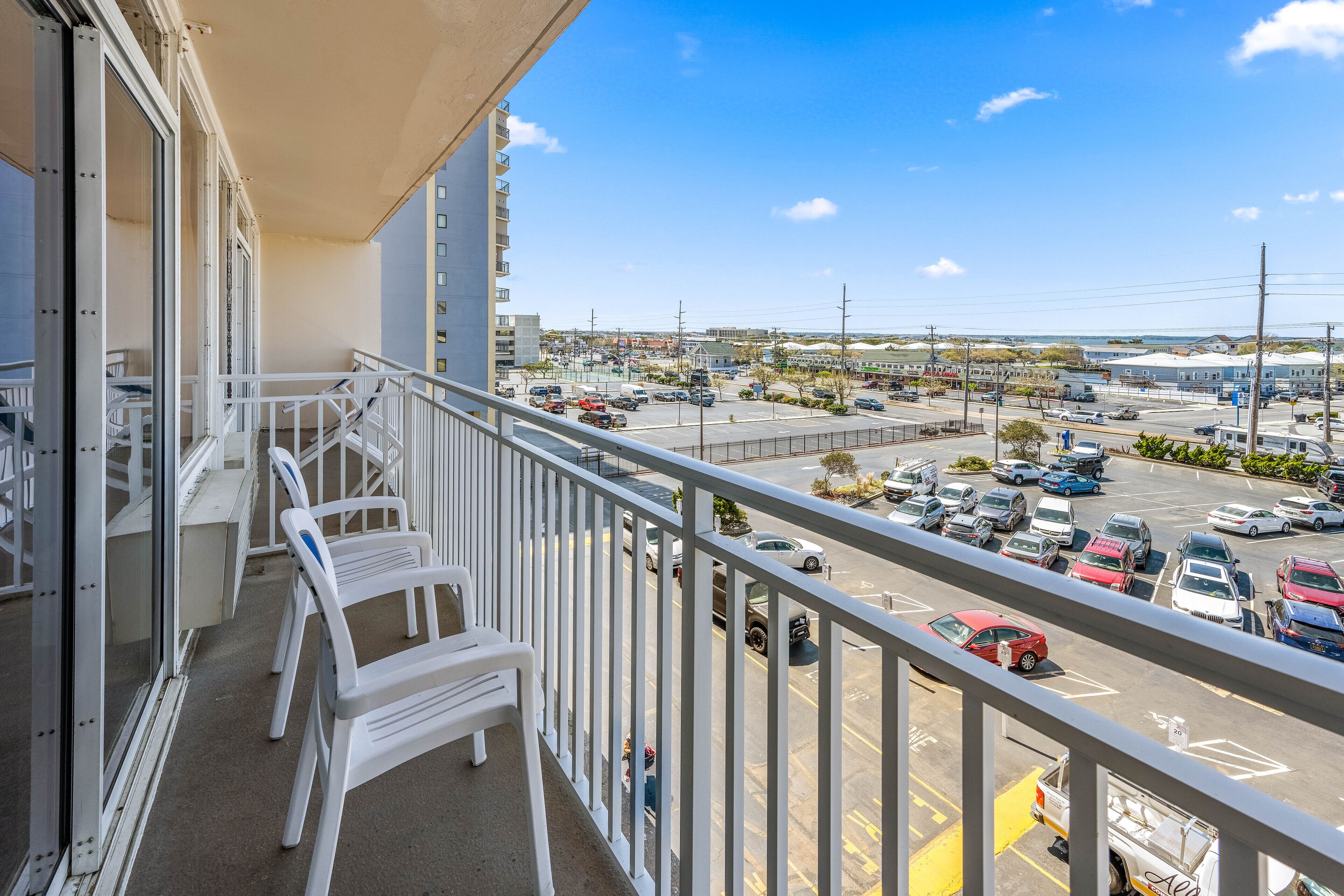 A balcony with white railings, a view of a parking lot, and a distant cityscape.