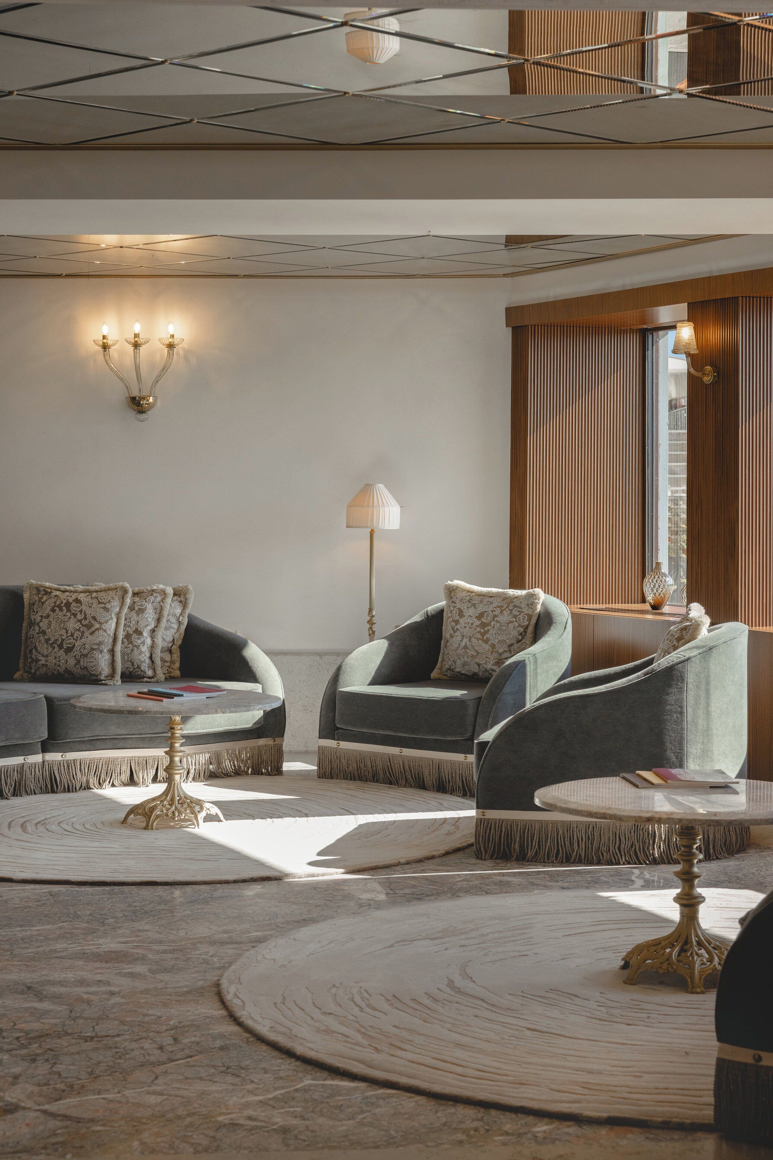 A modern living room with a sofa, armchairs, and a coffee table, featuring a geometric ceiling design and a large window with vertical blinds.