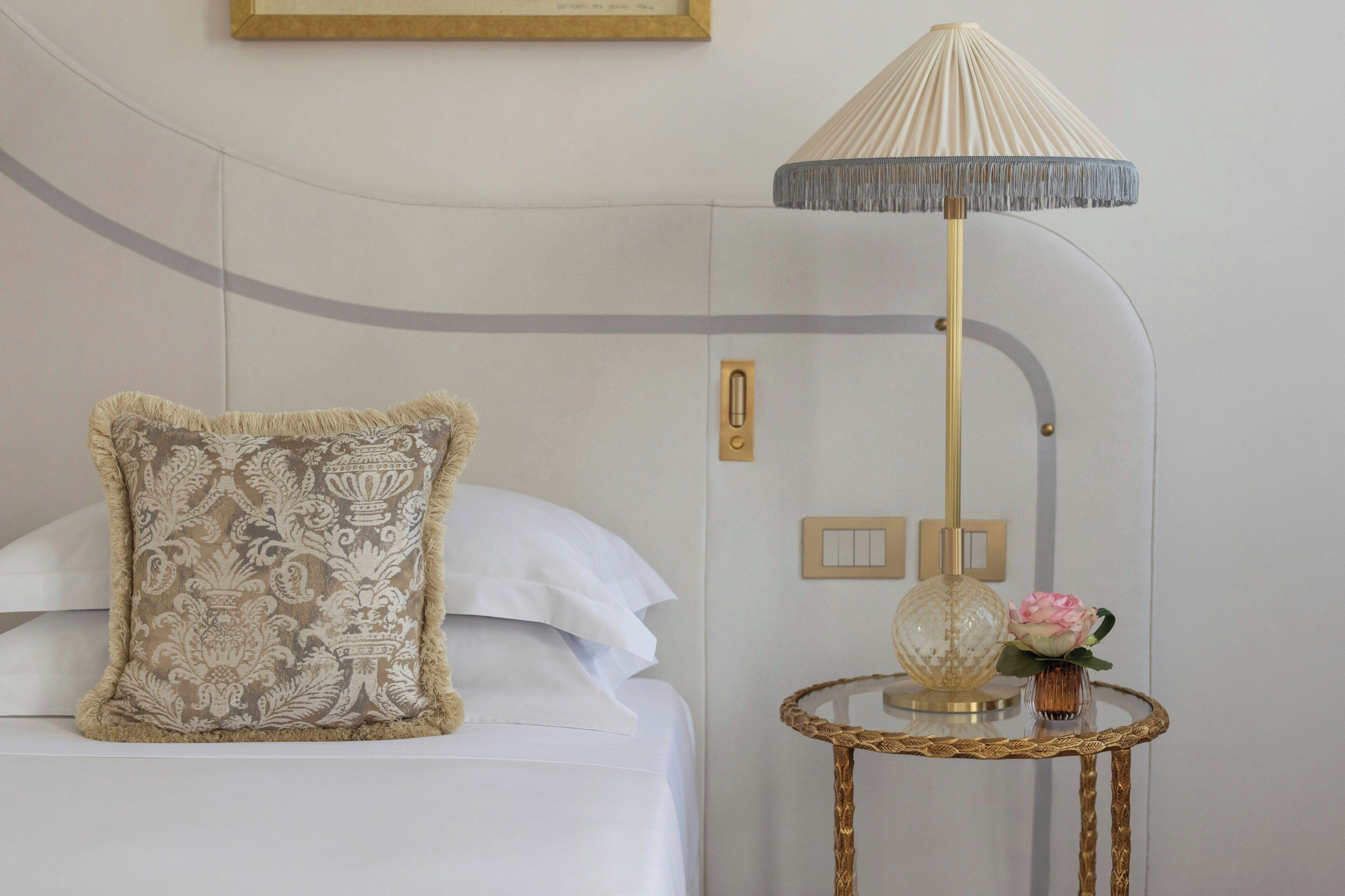 Un lit bien fait, agrémenté d’un coussin décoratif, une lampe sur pied avec un abat-jour plissé et une petite table sur laquelle sont posés un vase et une fleur.