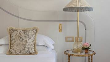 Un lit bien fait, agrémenté d’un coussin décoratif, une lampe sur pied avec un abat-jour plissé et une petite table sur laquelle sont posés un vase et une fleur.