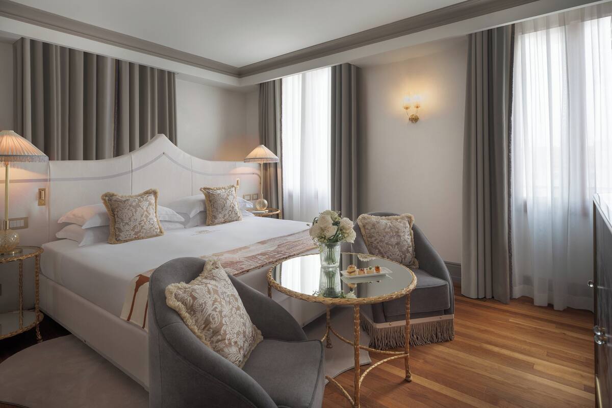 Une chambre d’hôtel avec un grand lit, deux fauteuils, une petite table ronde et un vase de fleurs.