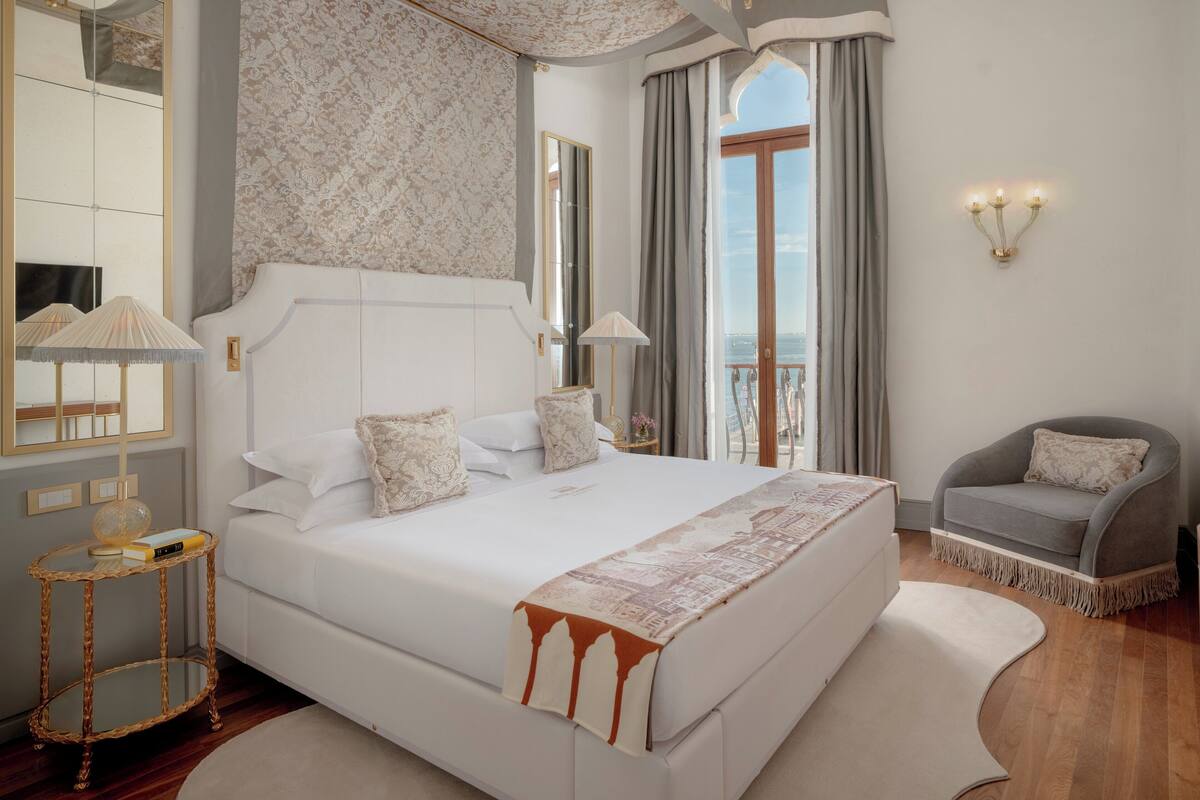 Une chambre spacieuse avec un grand lit, un fauteuil gris, un miroir et une vue sur l’extérieur.