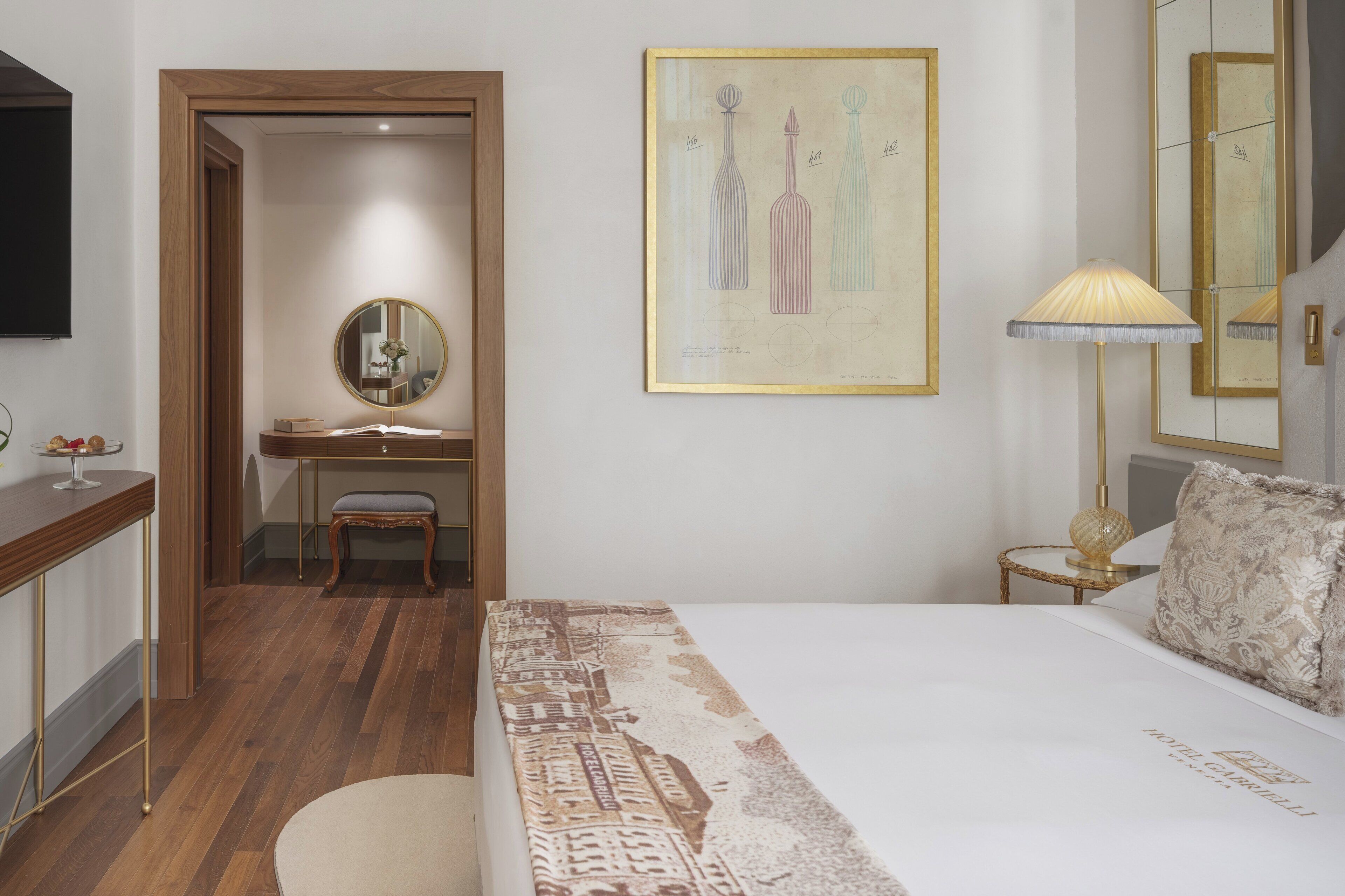 A hotel room with a bed, a desk, a chair, a mirror, and a wall-mounted lamp.