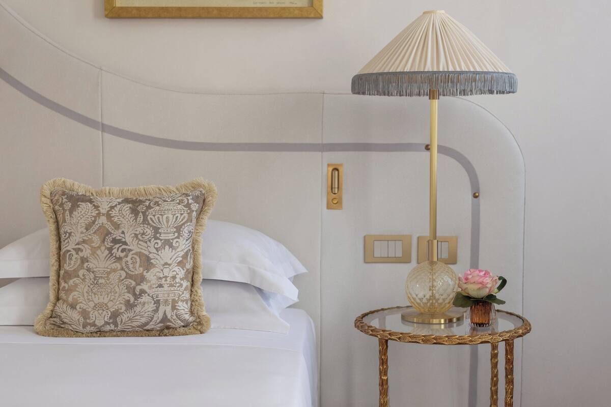 Un lit bien fait, agrémenté d’un coussin décoratif, une lampe avec un abat-jour à franges et une petite table sur laquelle est posé un vase de fleurs.