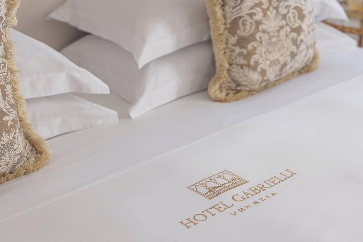 Un lit d’hôtel avec du linge de lit blanc et un logo indiquant « HÔTEL GABRIELI VENEZIA ».