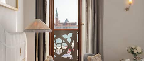 Habitación de hotel con cama, una silla, una lámpara y vista a un paisaje urbano a través de una ventana con cortinas.