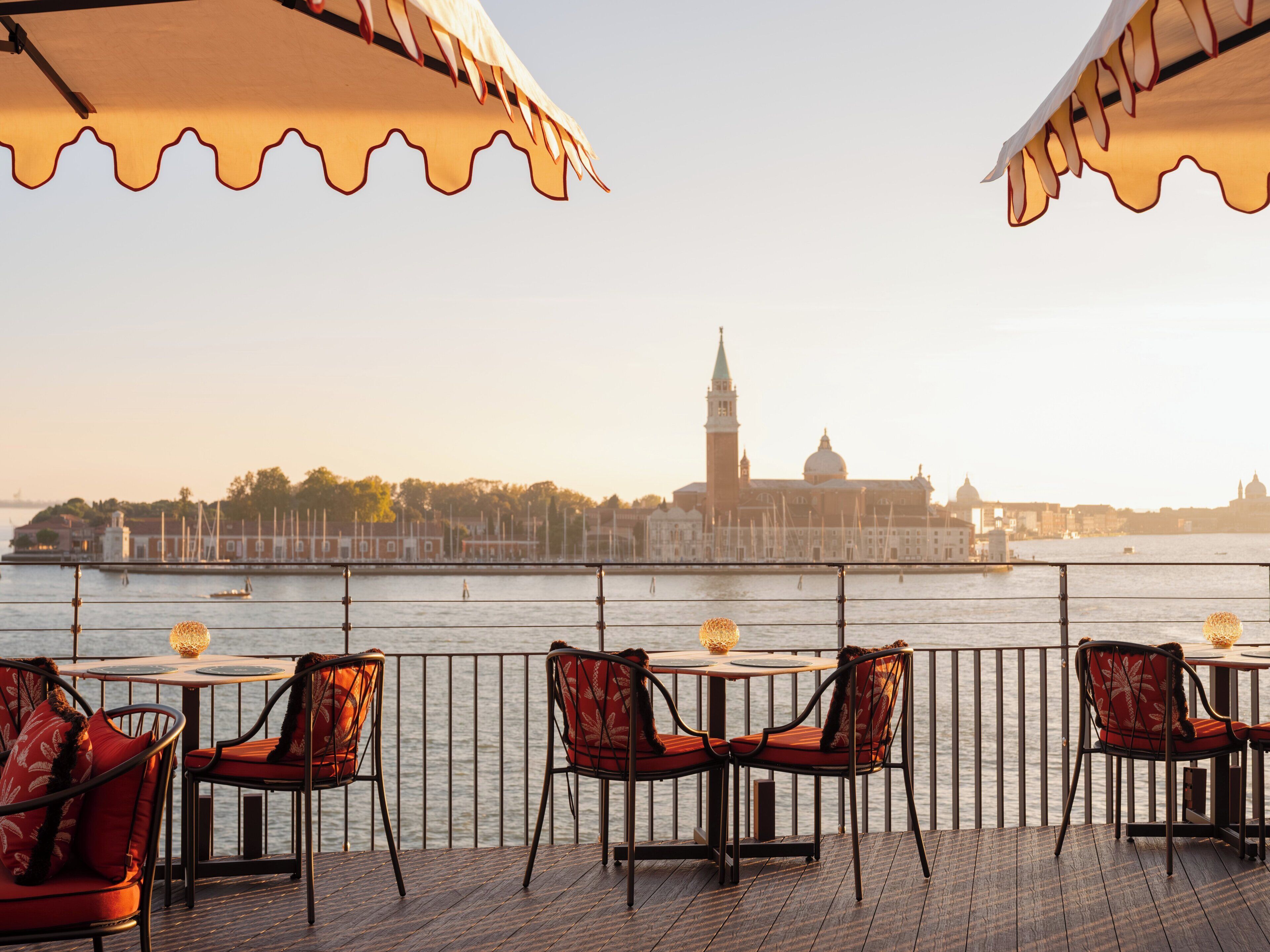 A waterfront terrace with tables and chairs, overlooking a river with buildings in the background.