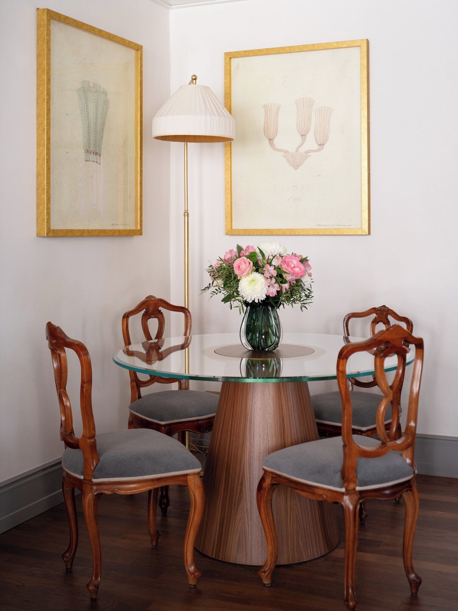 A dining area with a round glass table surrounded by wooden chairs with cushioned seats. There is a vase with flowers on the table, and two framed artworks on the wall.