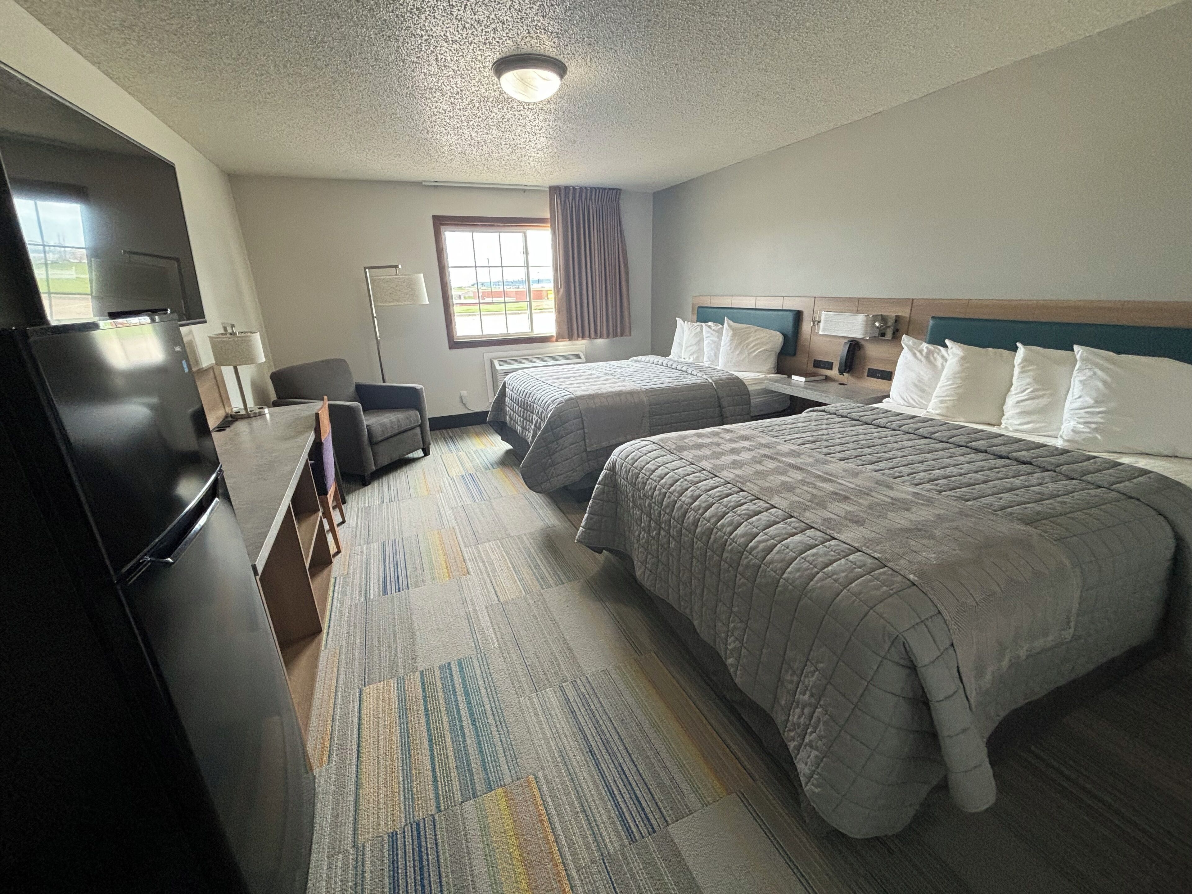 A hotel room with two beds, a desk, a refrigerator, and a window with curtains.