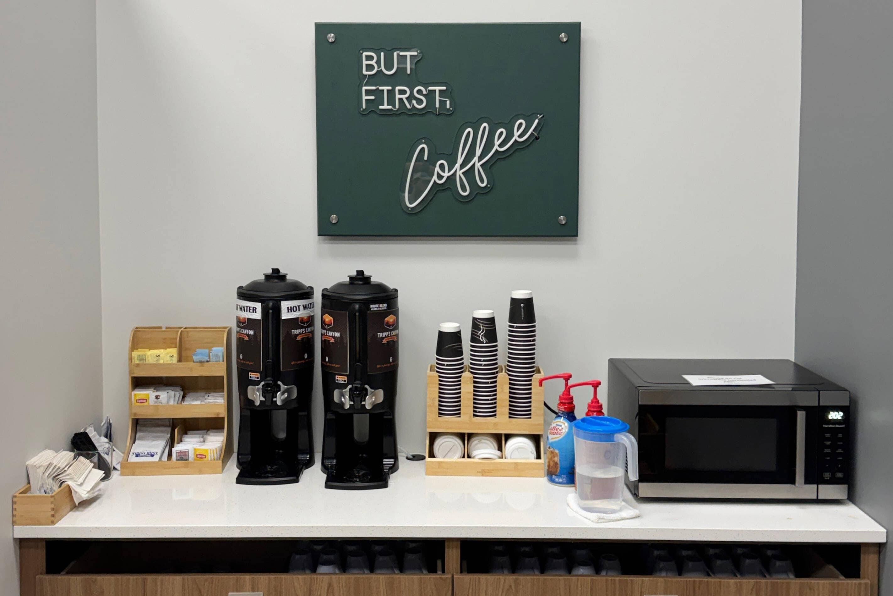Un coin café équipé de deux machines à café, d’une pile de gobelets jetables, d’un four à micro-ondes et d’un distributeur d’eau.