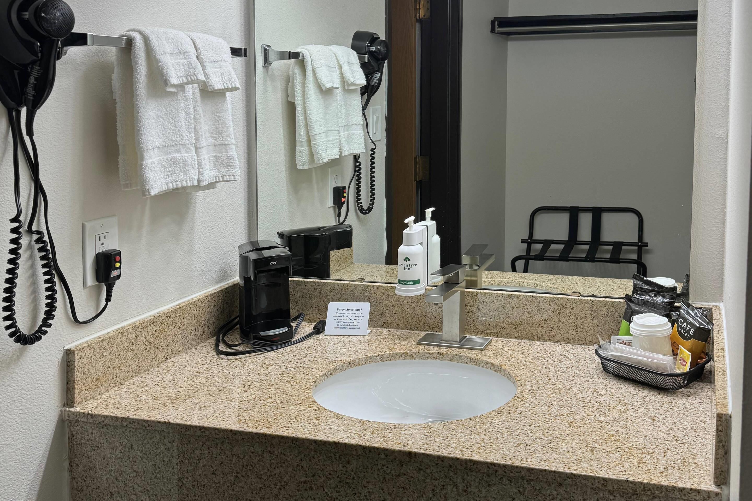 A hotel bathroom with a granite countertop, a sink, a mirror, a hairdryer, a towel rack, a coffee maker, and toiletries.
