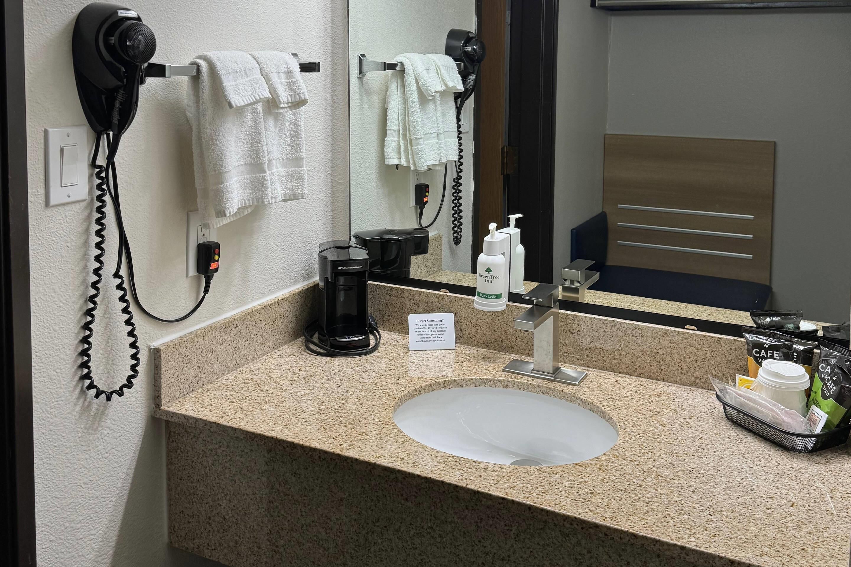 Baño de hotel con encimera de granito, lavabo, espejo, secador de pelo, toallero y cafetera.