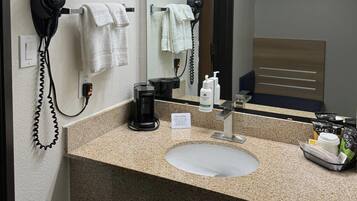 Baño de hotel con encimera de granito, lavabo, espejo, secador de pelo, toallero y cafetera.
