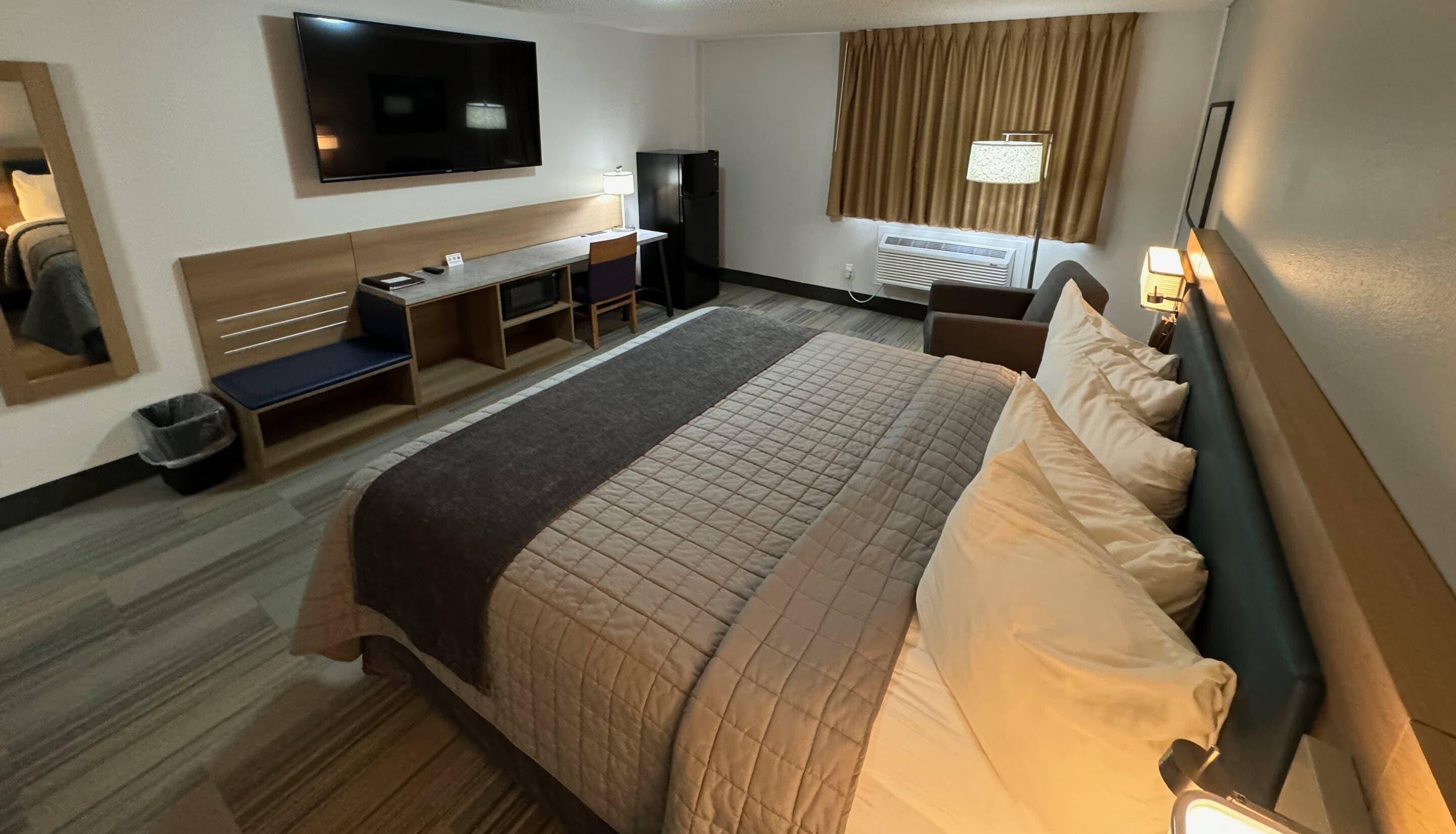 A hotel room with a large bed, a desk with a chair, a television, an air conditioning unit, and a window with curtains.