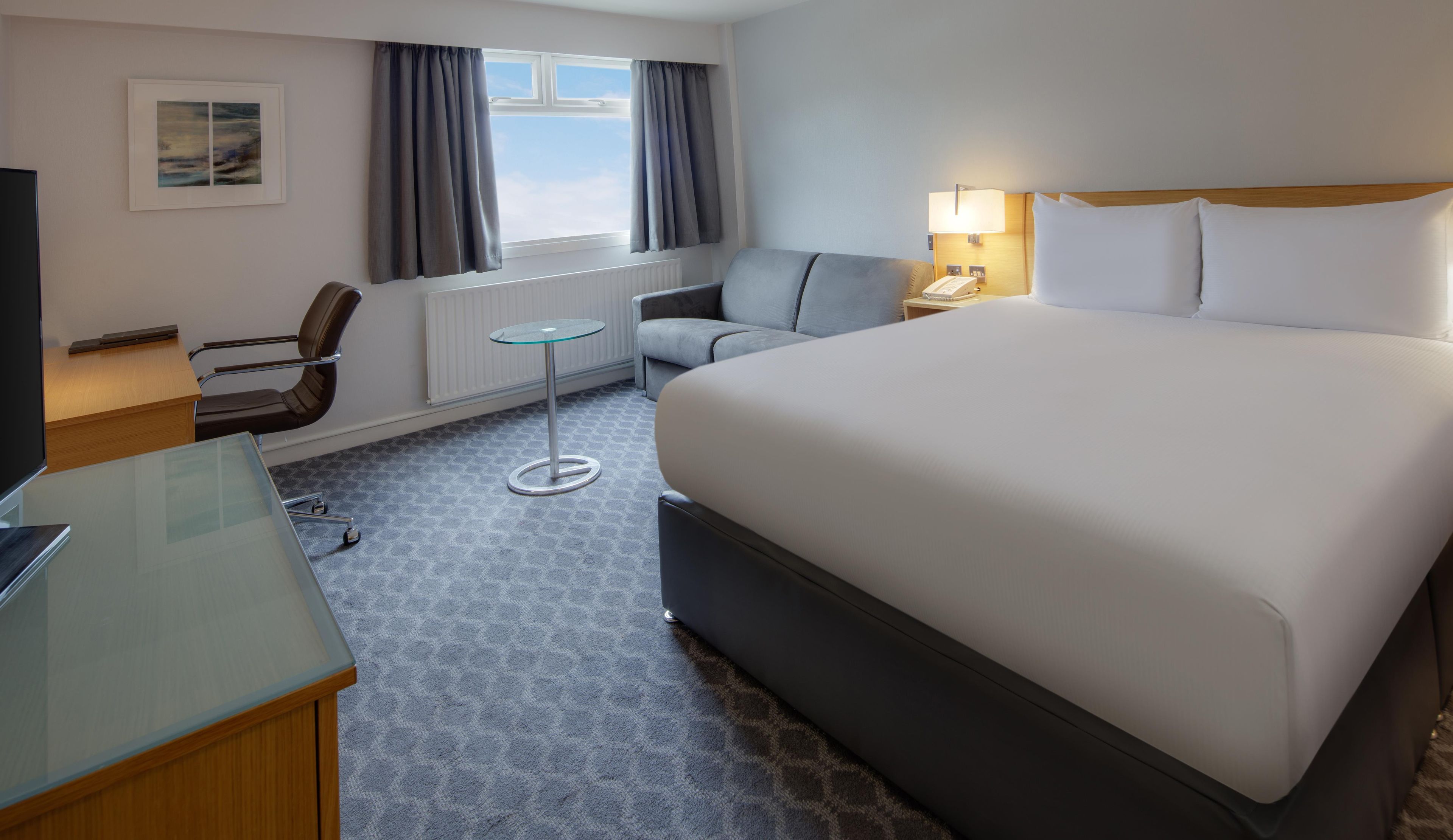 Habitación de hotel con cama, escritorio, silla, mesita y ventana con cortinas.