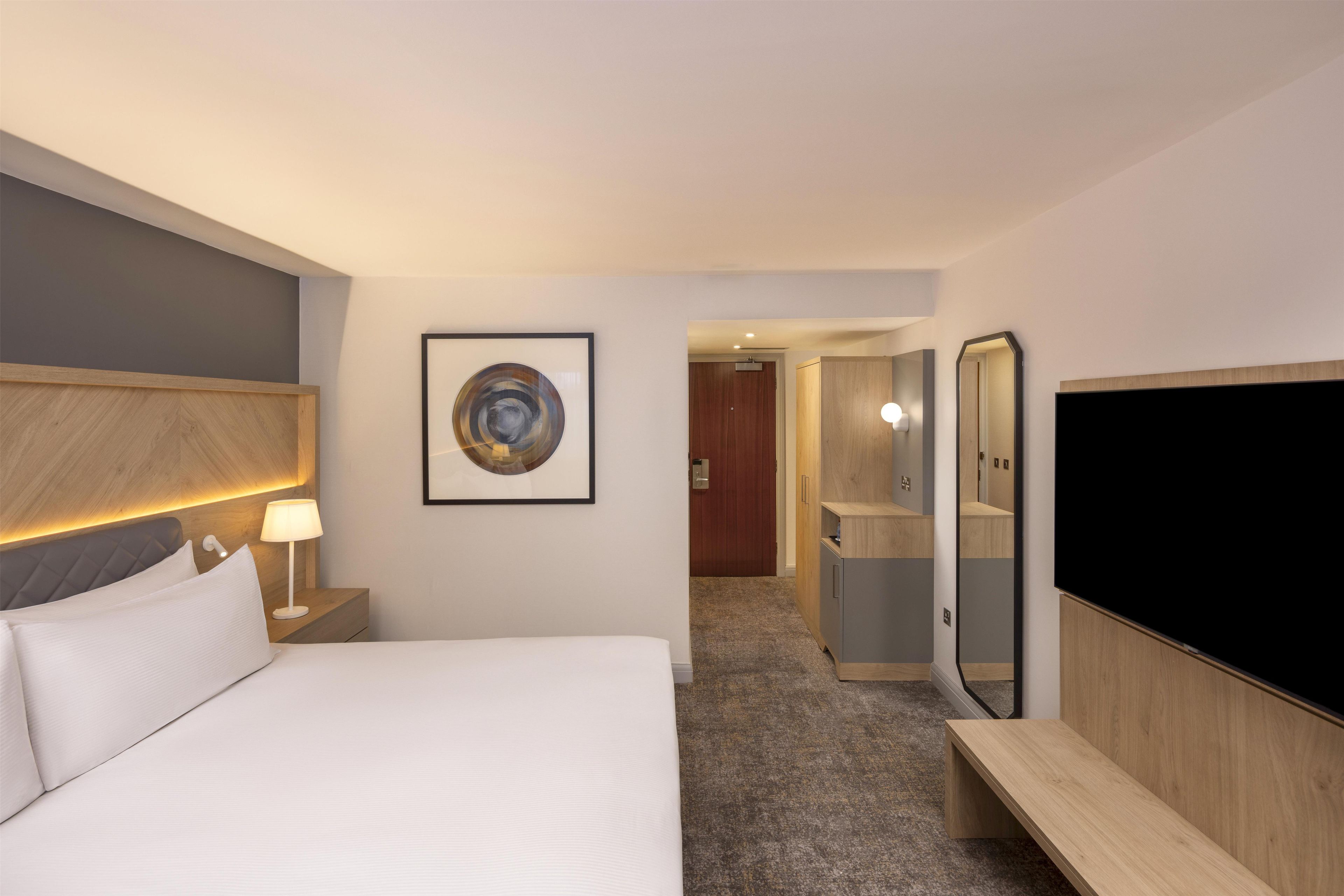 Una habitación de hotel con cama, lámparas de noche, una obra de arte enmarcada, un espejo y un televisor.