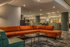 A modern hotel lobby with a sectional sofa, a coffee table, and a bar area in the background.