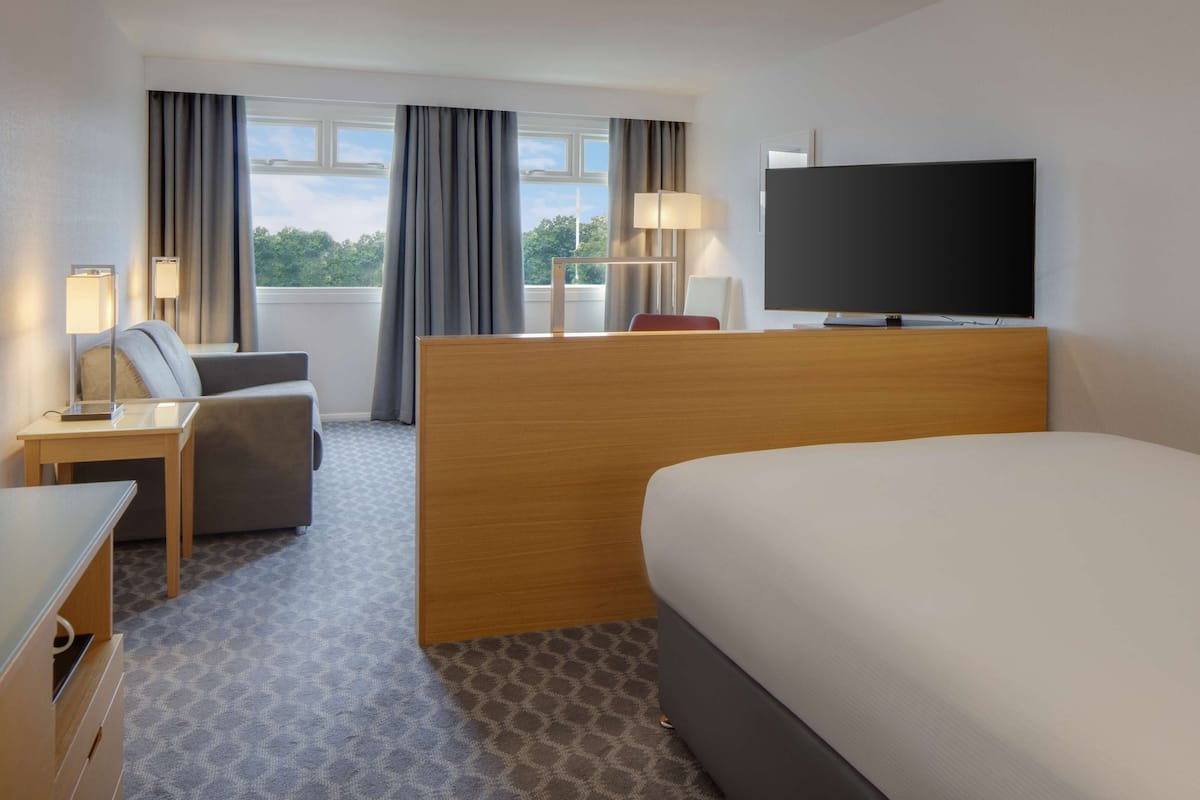 A hotel room with a bed, a desk with a lamp, a chair, a TV, and a window with curtains.
