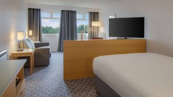 Una habitación de hotel con una cama, un escritorio con lámpara, una silla, un televisor y un ventanal con cortinas.
