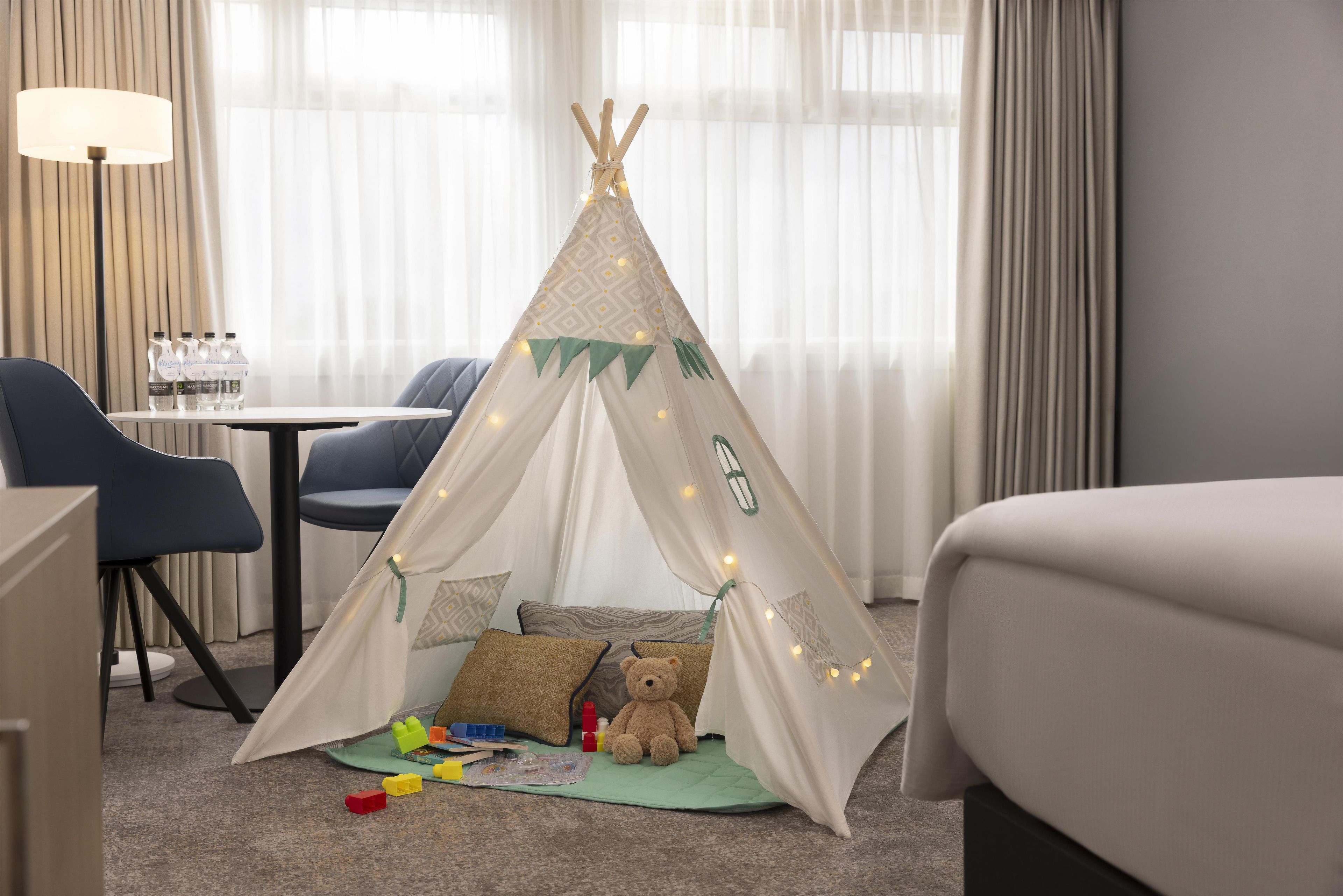 Habitación de hotel con una tienda tipi, una lámpara, una mesa con botellas de agua y un osito de peluche.