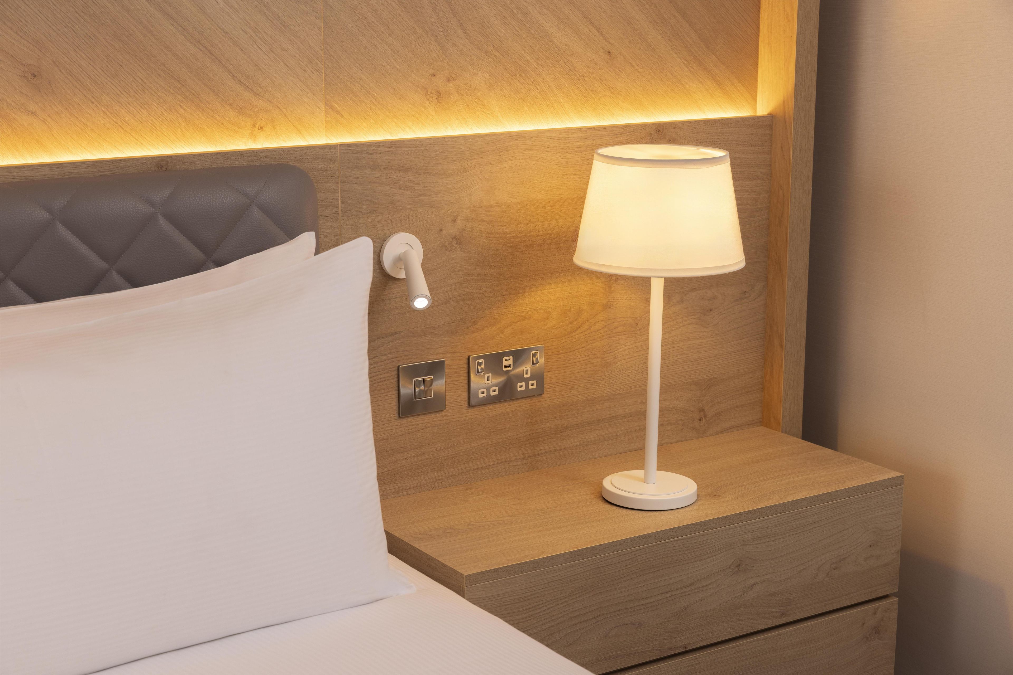 Una cama bien hecha con almohada, una lámpara de noche y un interruptor de luz en la pared.