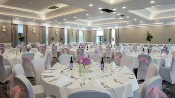 A large banquet hall with round tables set for a formal event, featuring white tablecloths, neatly arranged cutlery, and floral centerpieces.