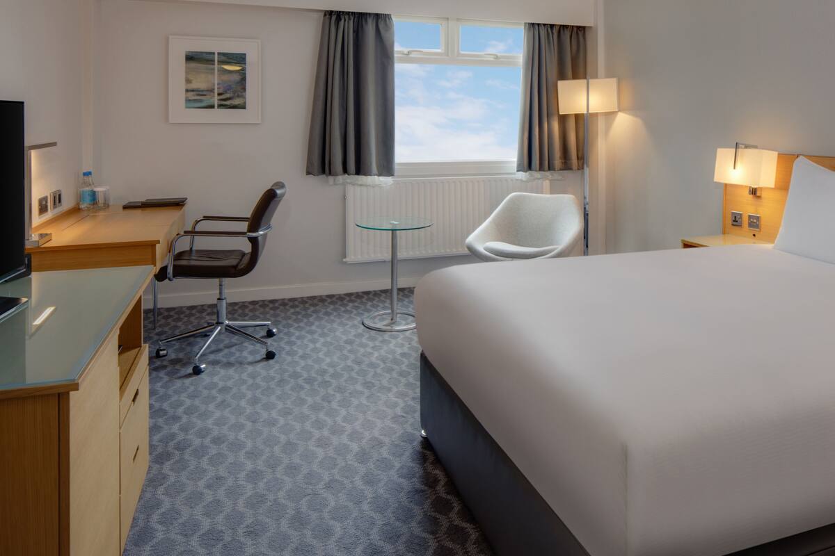 A hotel room with a bed, a desk with a chair, a small table, and a window with curtains.