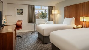 Ein Hotelzimmer mit zwei Betten, einem Schreibtisch, einem Stuhl, einem kleinen Tisch und einem Fenster mit Vorhängen.