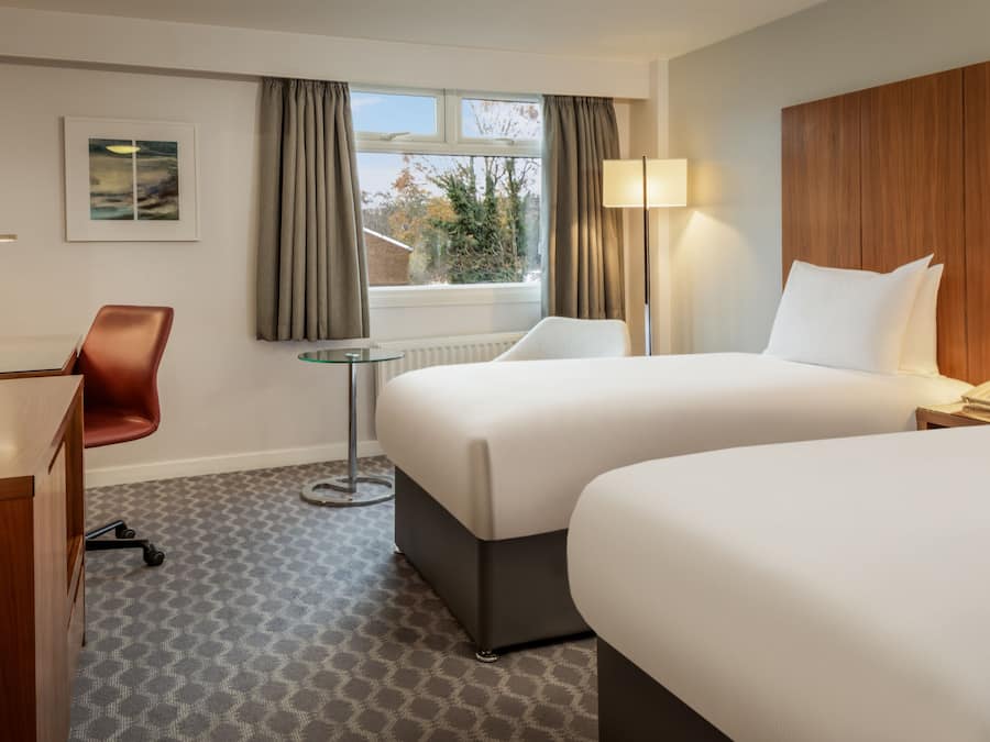 A hotel room with two beds, a desk, a chair, a small table, and a window with curtains.