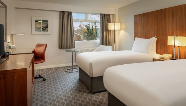 A hotel room with two beds, a desk, a chair, a small table, and a window with curtains. - Hilton London Watford (Watford)