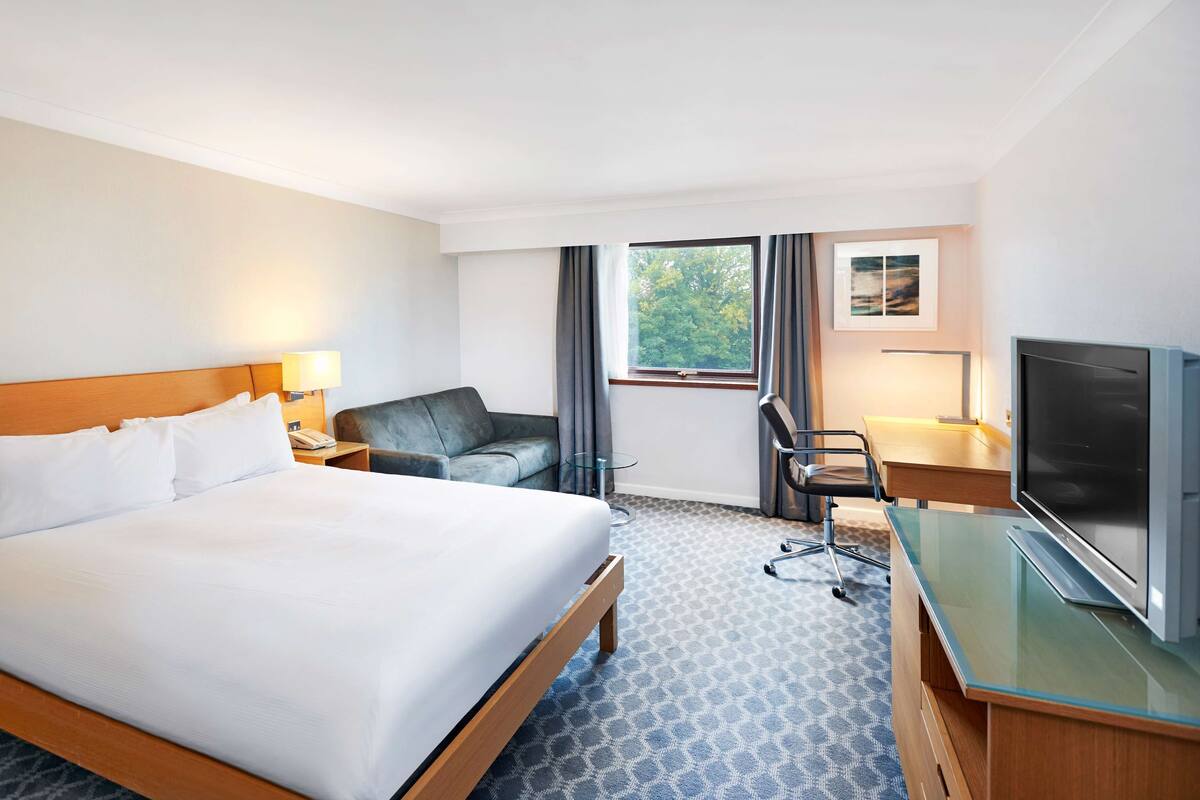 A hotel room with a large bed, a desk with a TV, a chair, a small table, and a window with curtains.