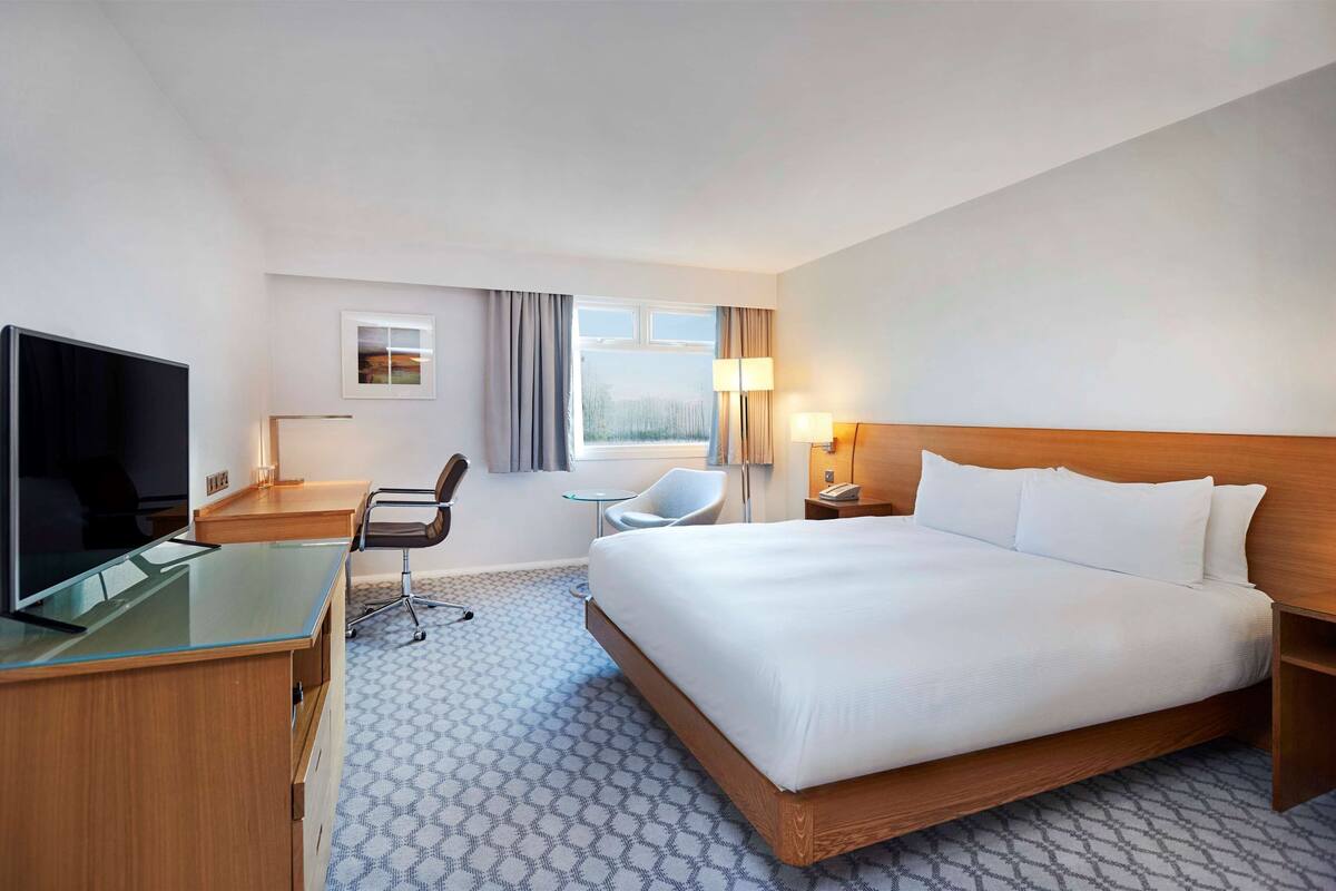 A hotel room with a large bed, a desk with a computer, a chair, a TV, and a window with curtains.